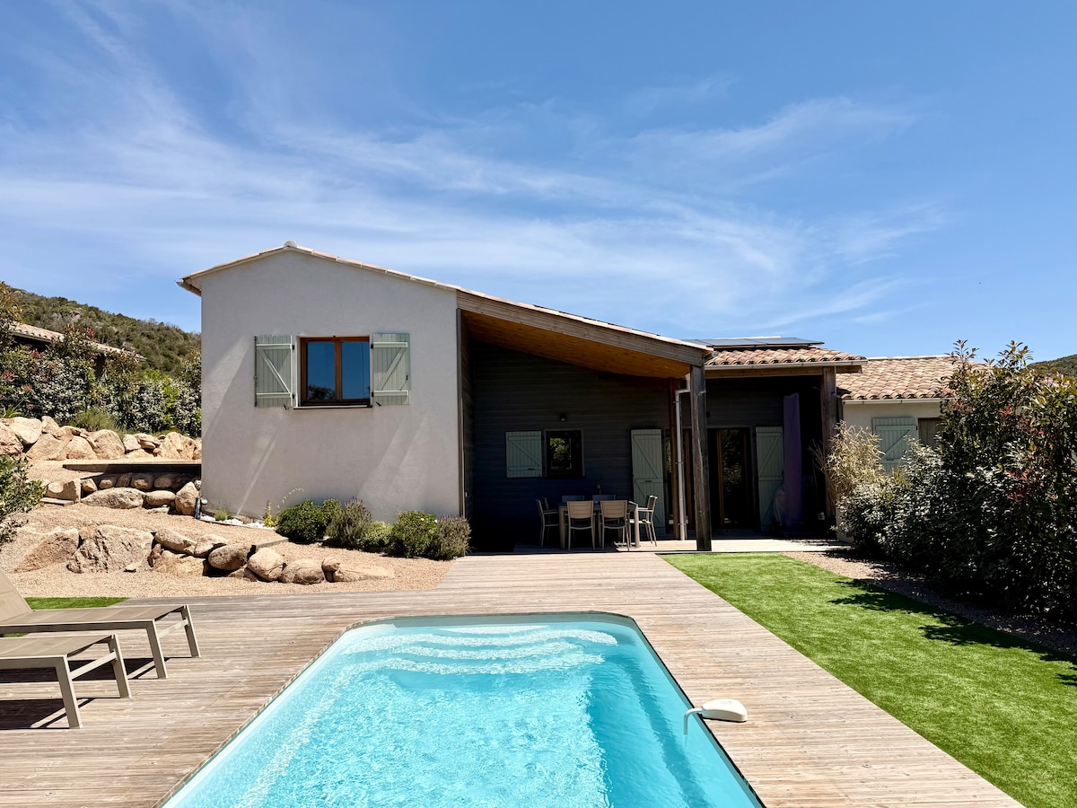 The exterior of the house features a private heated pool and a spacious deck area. A lush green lawn surrounds the terrace, with outdoor seating visible. The facade reflects a harmonious blend of modern and traditional elements under a clear blue sky.