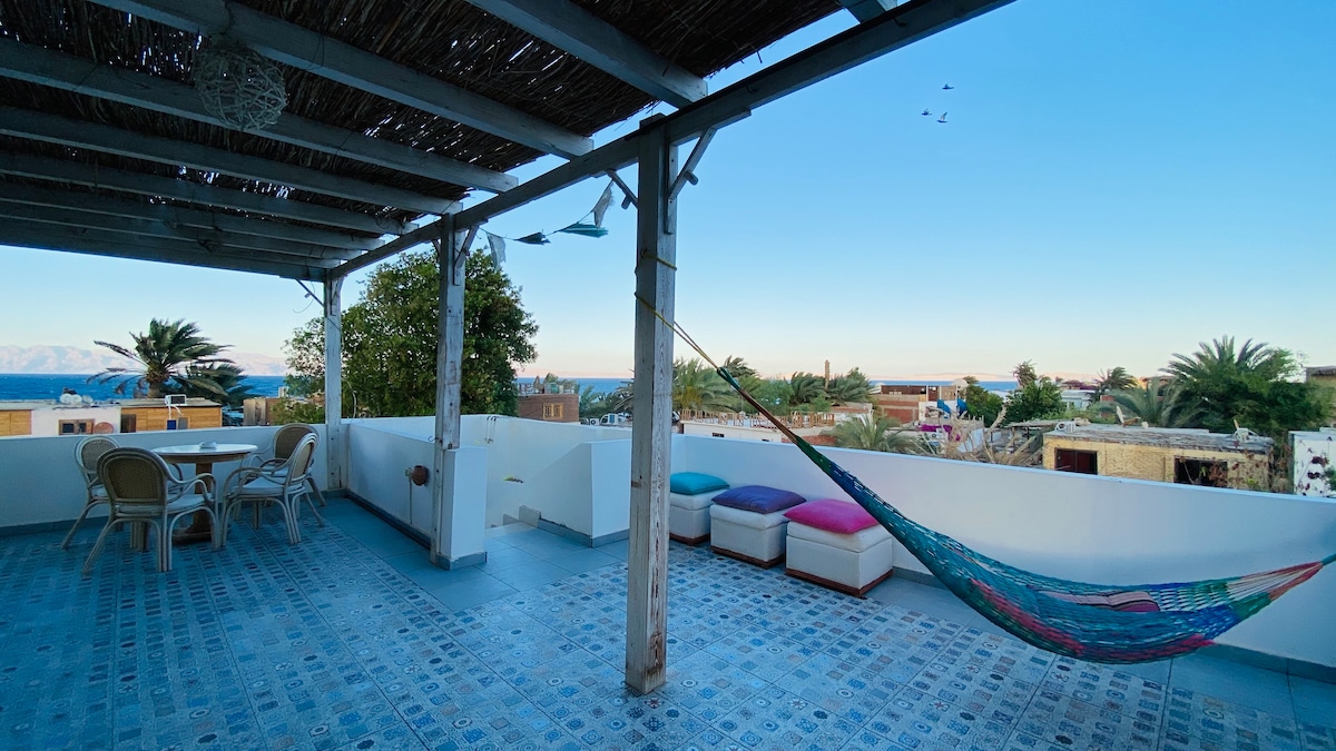 A spacious rooftop area features comfortable seating arrangements, including a dining table with chairs. A colorful hammock is hung between posts, inviting relaxation. The blue-tiled flooring enhances the serene atmosphere, while a panoramic view showcases nearby palm trees and the sea in the distance.