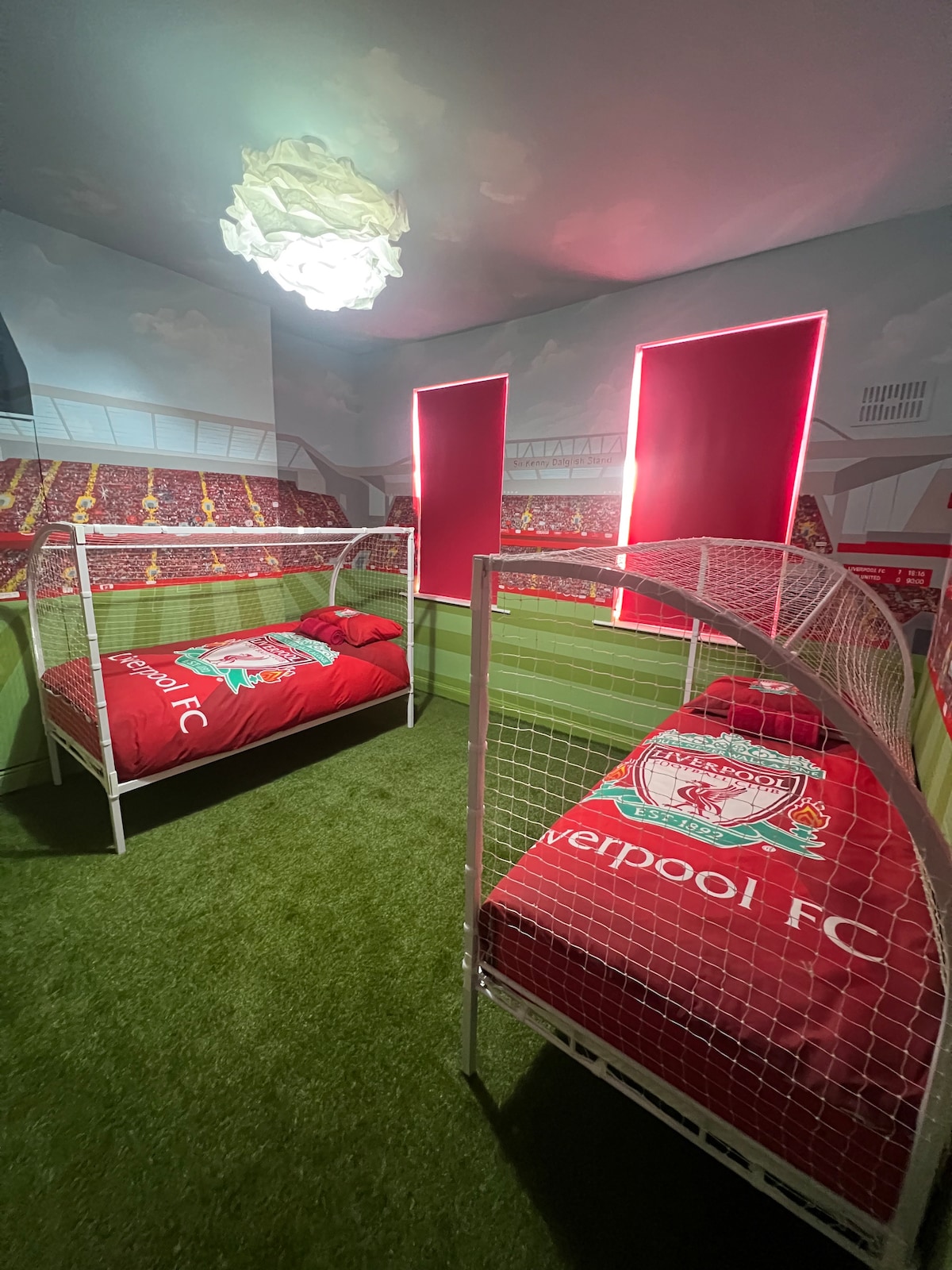 A themed room features two beds designed to resemble goal nets, adorned with Liverpool FC bedding. The walls depict a stadium backdrop, while bright red curtains frame two windows. A textured green carpet covers the floor, contributing to a sports-oriented atmosphere.