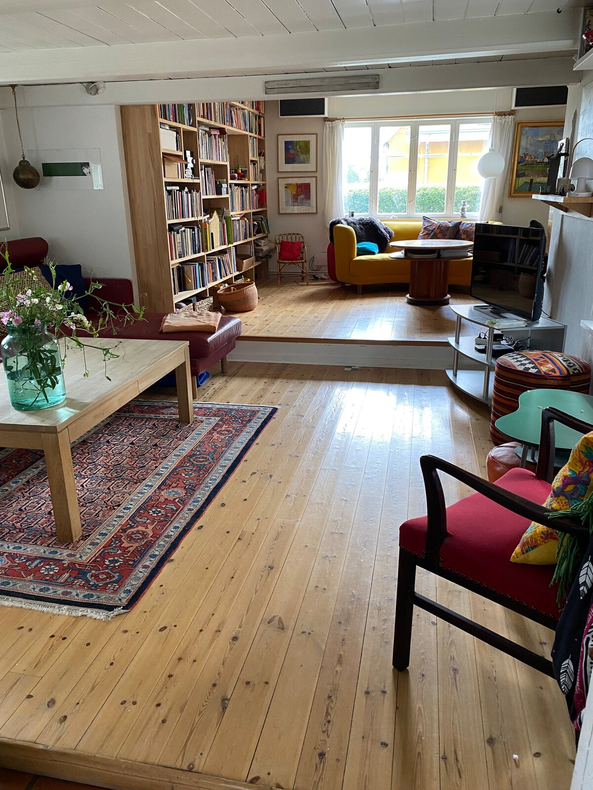 A cozy living area features wooden flooring and a large coffee table at its center. Shelves filled with books line one wall, while colorful seating options create an inviting atmosphere. Natural light fills the space through windows, offering views of the garden outside.