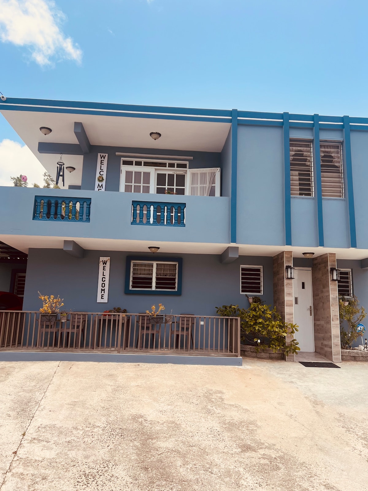 The exterior of a two-story building is showcased, featuring a blue-painted facade with white accents. A welcoming sign is displayed prominently. The entrance is flanked by neatly arranged plants, and balconies are visible on the upper level, enhancing the overall inviting appearance.