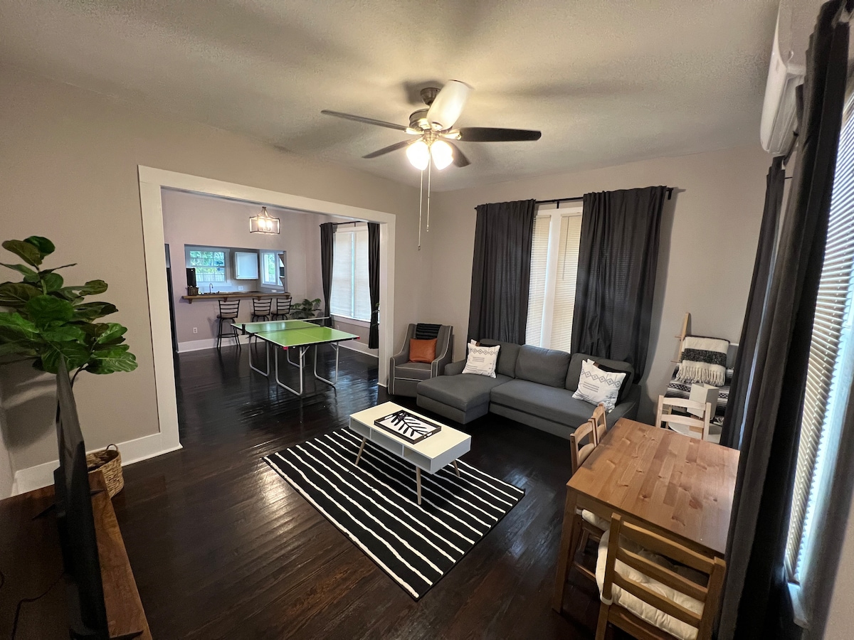 A spacious living area features a sectional sofa and a coffee table, with a ceiling fan overhead. A dining area is visible through an open entryway. Large windows are covered by dark curtains, allowing natural light to filter in. A playful area rug adds contrast to the dark wooden flooring.