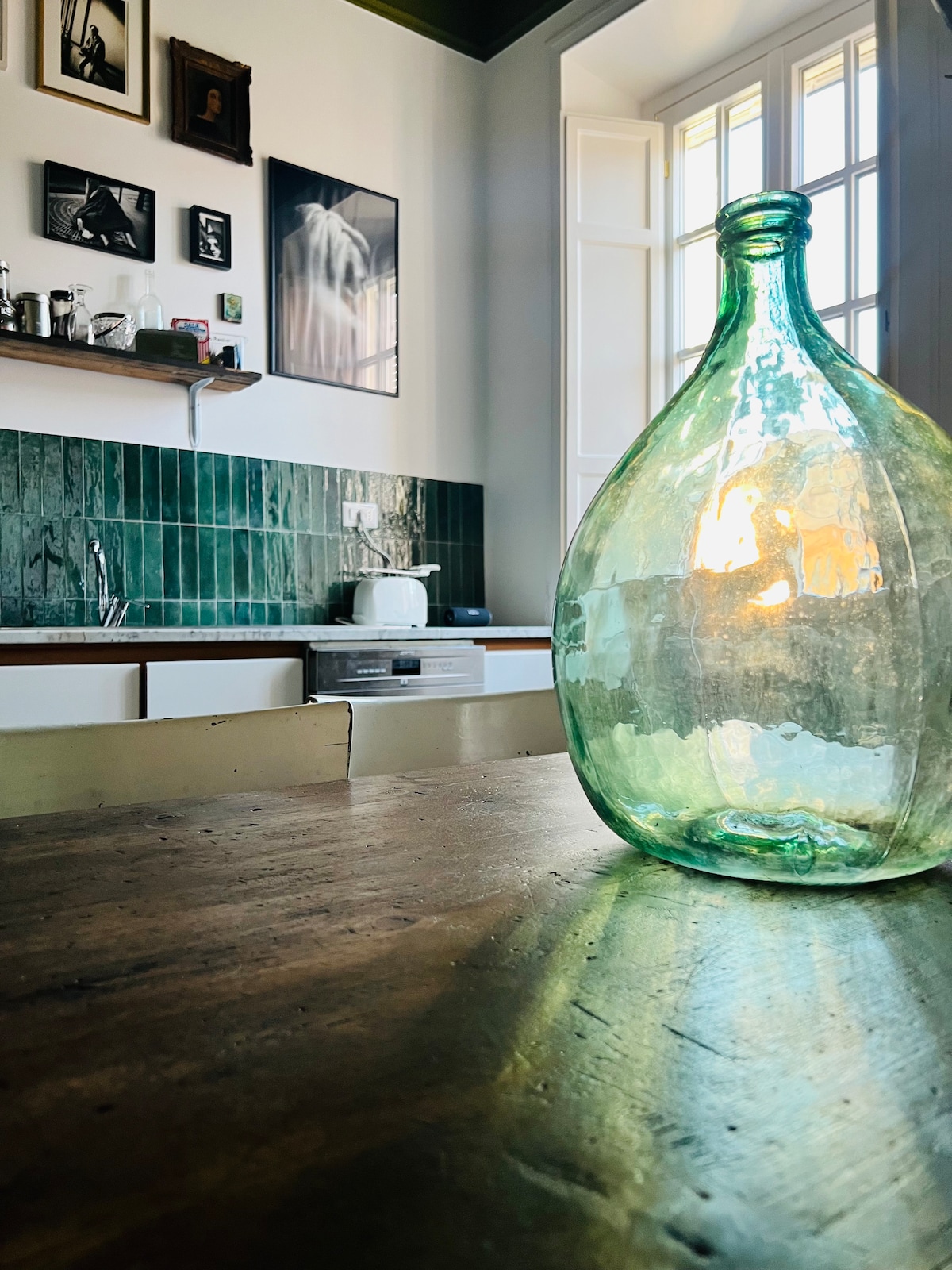 A vintage green glass bottle sits on a wooden table, capturing the warm glow of light. Behind it, a kitchen with green tile backsplash and wooden shelves displays various kitchen items, including a toaster, creating a cozy atmosphere.