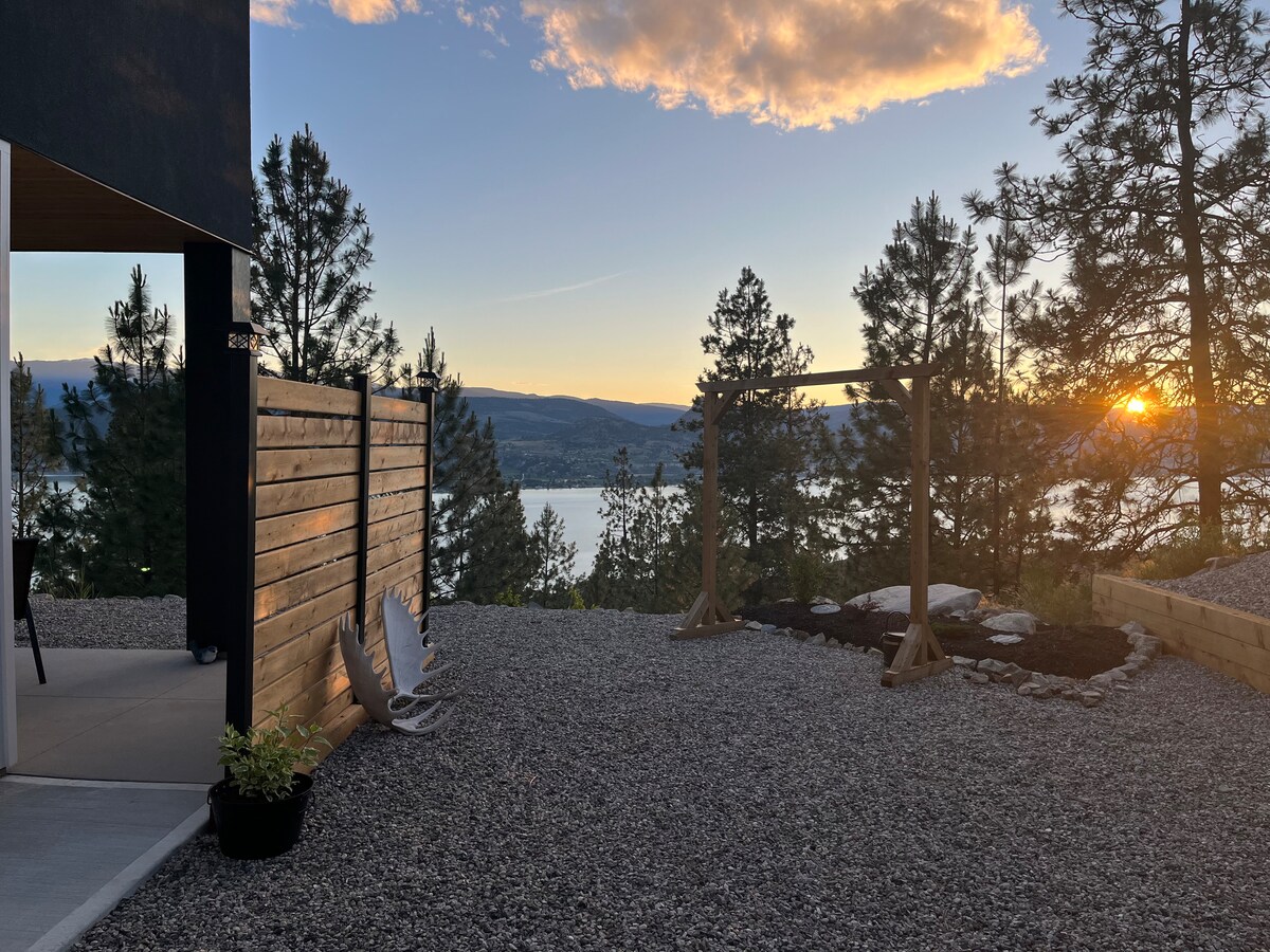 A gravel outdoor space is presented, featuring two lounge chairs positioned to enjoy views of Okanagan Lake and surrounding trees. The setting sun casts a warm glow, enhancing the serene atmosphere, while a wooden swing structure is visible in the background.