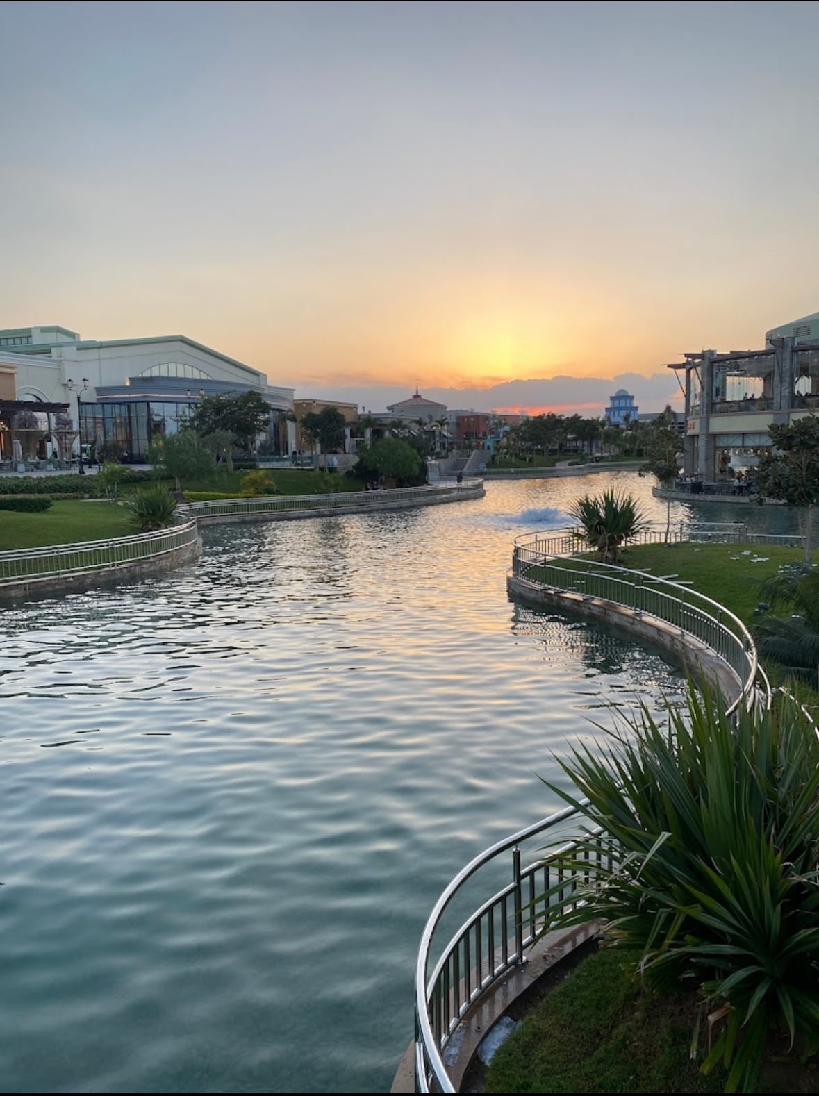 A serene waterway winds through well-maintained greenery, with gentle curves reflecting the soft glow of sunset. Light dances on the water's surface, surrounded by landscaped gardens, creating a tranquil atmosphere. The backdrop features modern buildings framed by a clear sky transitioning to twilight.