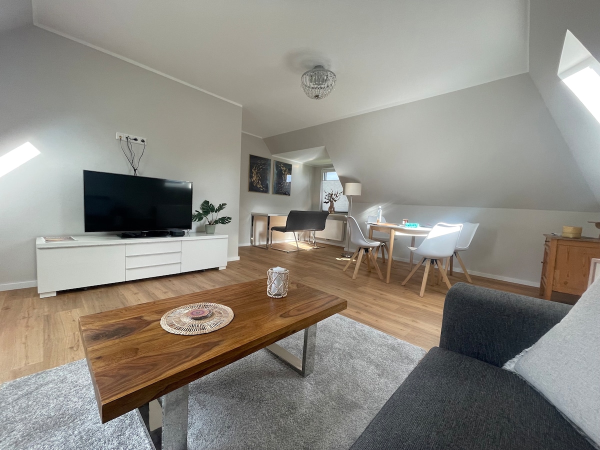 A spacious living area features a dark couch facing a wall-mounted television. Natural light enters through skylights, highlighting the light wood flooring. A wooden coffee table with a decorative centerpiece sits at the center, alongside a dining area with modern chairs and a small workspace in the background.