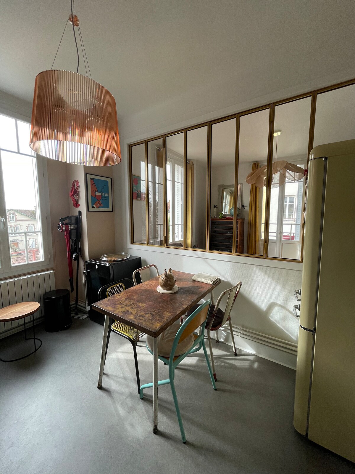 A dining area features a rustic table surrounded by several chairs, with a copper pendant light hanging above. A retro refrigerator is visible, along with a small side table. A window allows natural light to illuminate the space, complemented by gold-framed mirrors on the opposite wall.