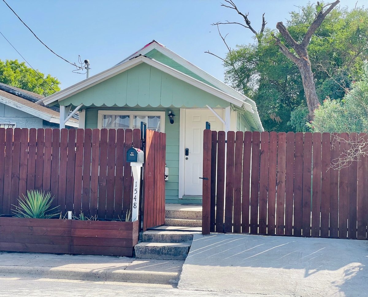 A charming green cottage is nestled behind a red wooden fence. The front door is framed by a small porch and is complemented by a mailbox positioned beside the walkway. Lush greenery is visible within the fenced area, contributing to the inviting exterior.