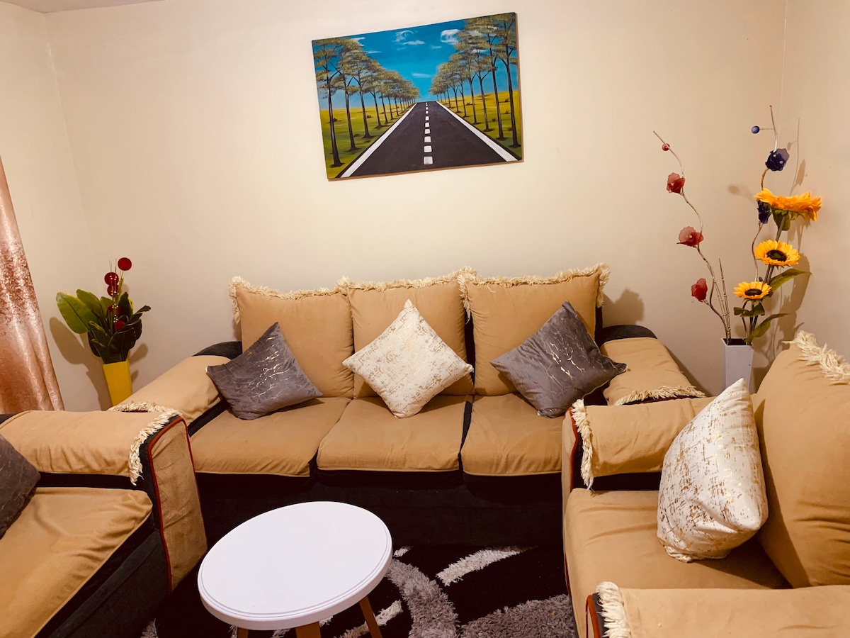 A comfortable seating area is furnished with a large, U-shaped sofa adorned with decorative cushions. A round coffee table is positioned in the center, complemented by a vibrant piece of wall art depicting a tree-lined road. Natural elements, including potted plants and flowers, add a touch of color.