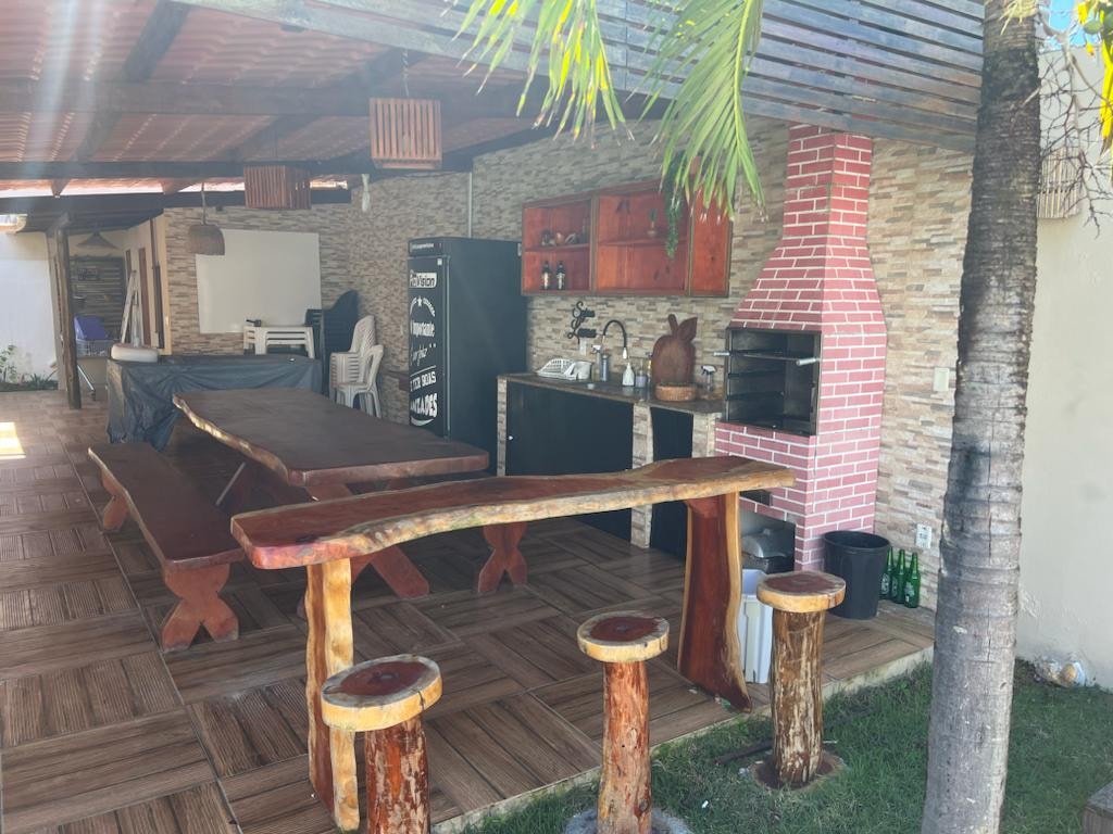 An outdoor leisure area features a spacious wooden dining table, surrounded by stools made from natural wood. A kitchenette with cabinetry is visible, along with a grill. The area is shaded by a pergola, and greenery enhances the inviting environment.