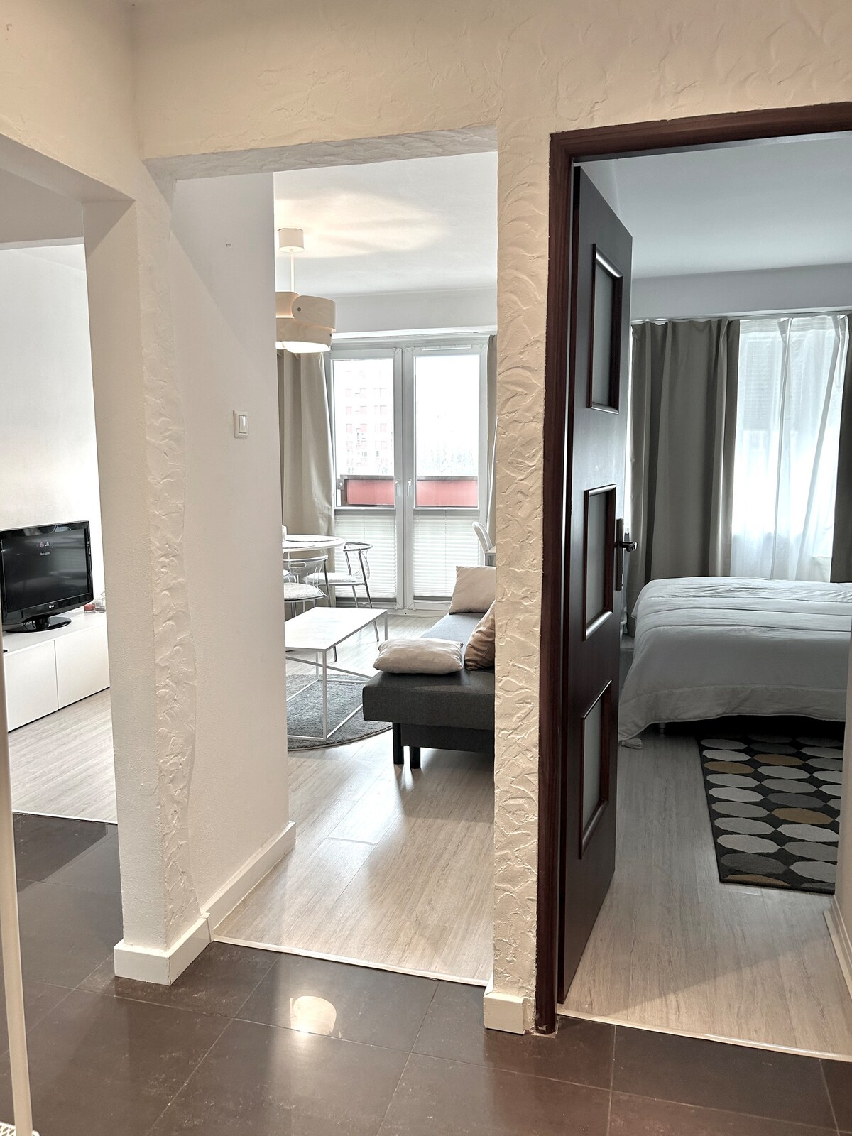 A view into the apartment reveals an open layout connecting the living area and bedroom. Natural light filters through large windows, highlighting a cozy seating arrangement and a spacious bed, framed by soft curtains. The room features neutral tones and modern furnishings.