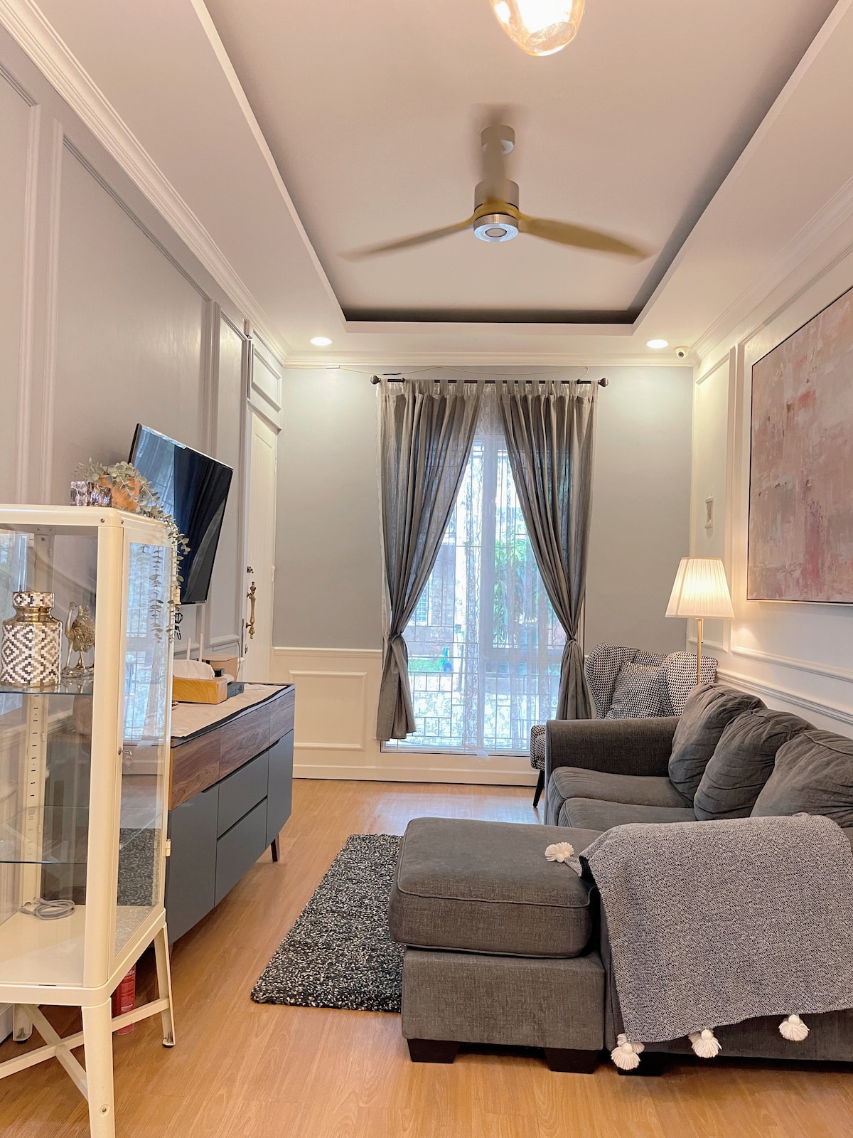 A comfortable living area is presented with a gray sectional sofa and an accent rug. Natural light enters through a tall window adorned with sheer curtains. A mounted television and decorative elements add to the modern style of the space.