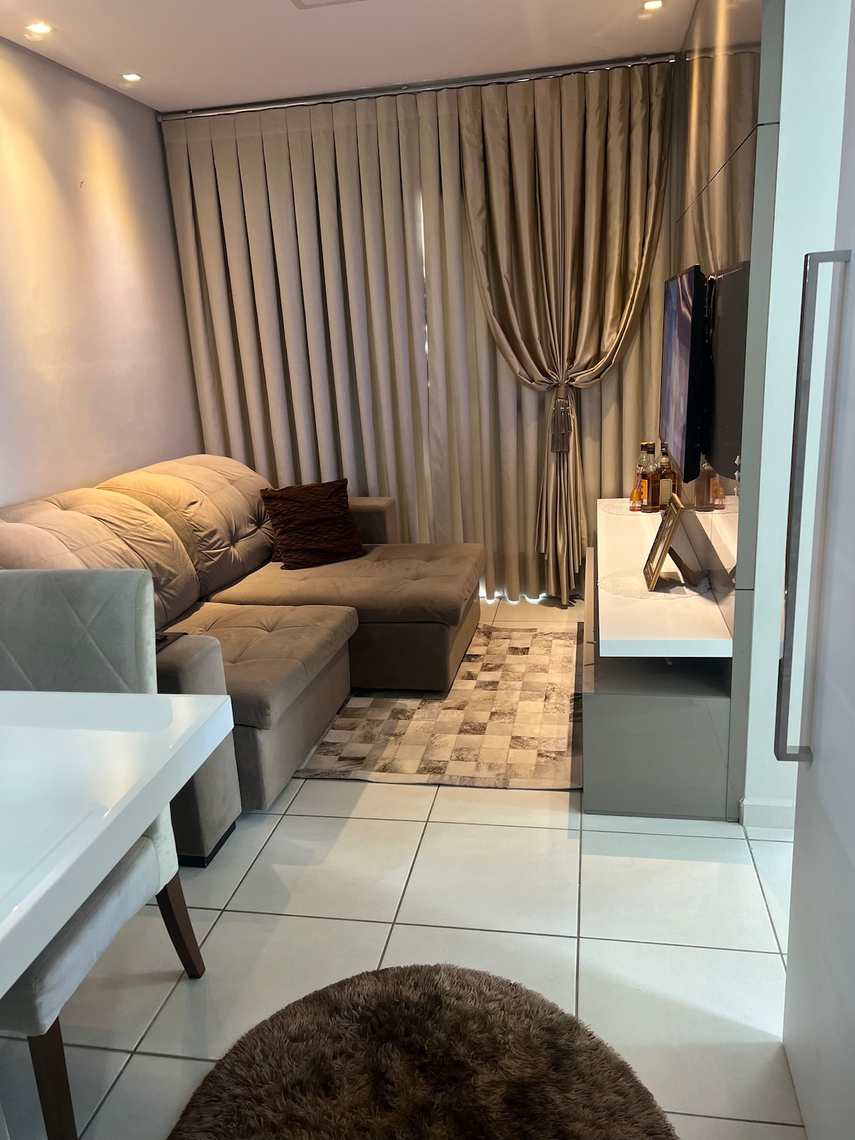 A compact living area is featured with a comfortable sectional sofa and a plush rug on tiled flooring. Elegant curtains frame a window, and a flat-screen television is mounted above a modern console table. A dining table with chairs is positioned nearby.
