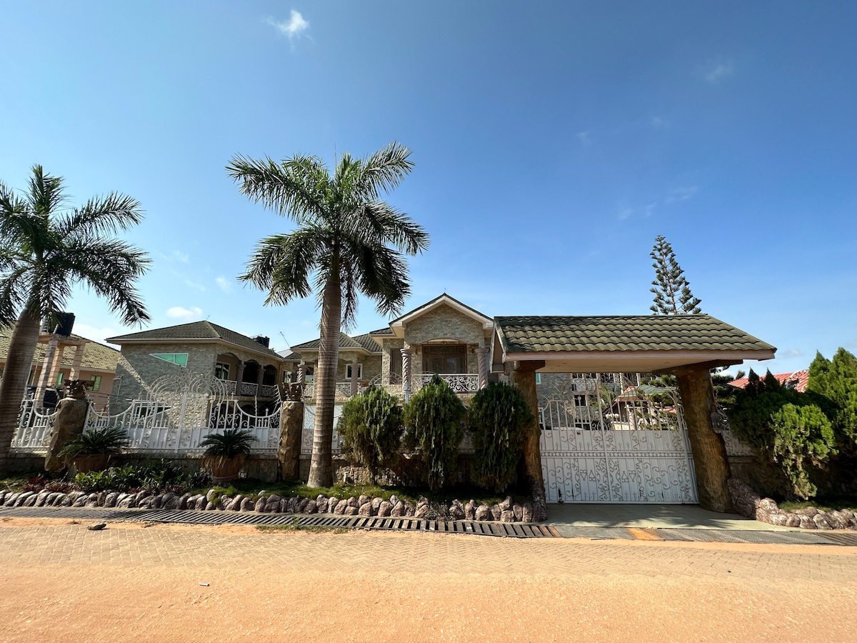 A grand mansion is framed by tall palm trees and lush greenery, with a decorative stone gate leading to the entrance. The detailed facade features stone accents and expansive windows, inviting natural light into the spacious interior.