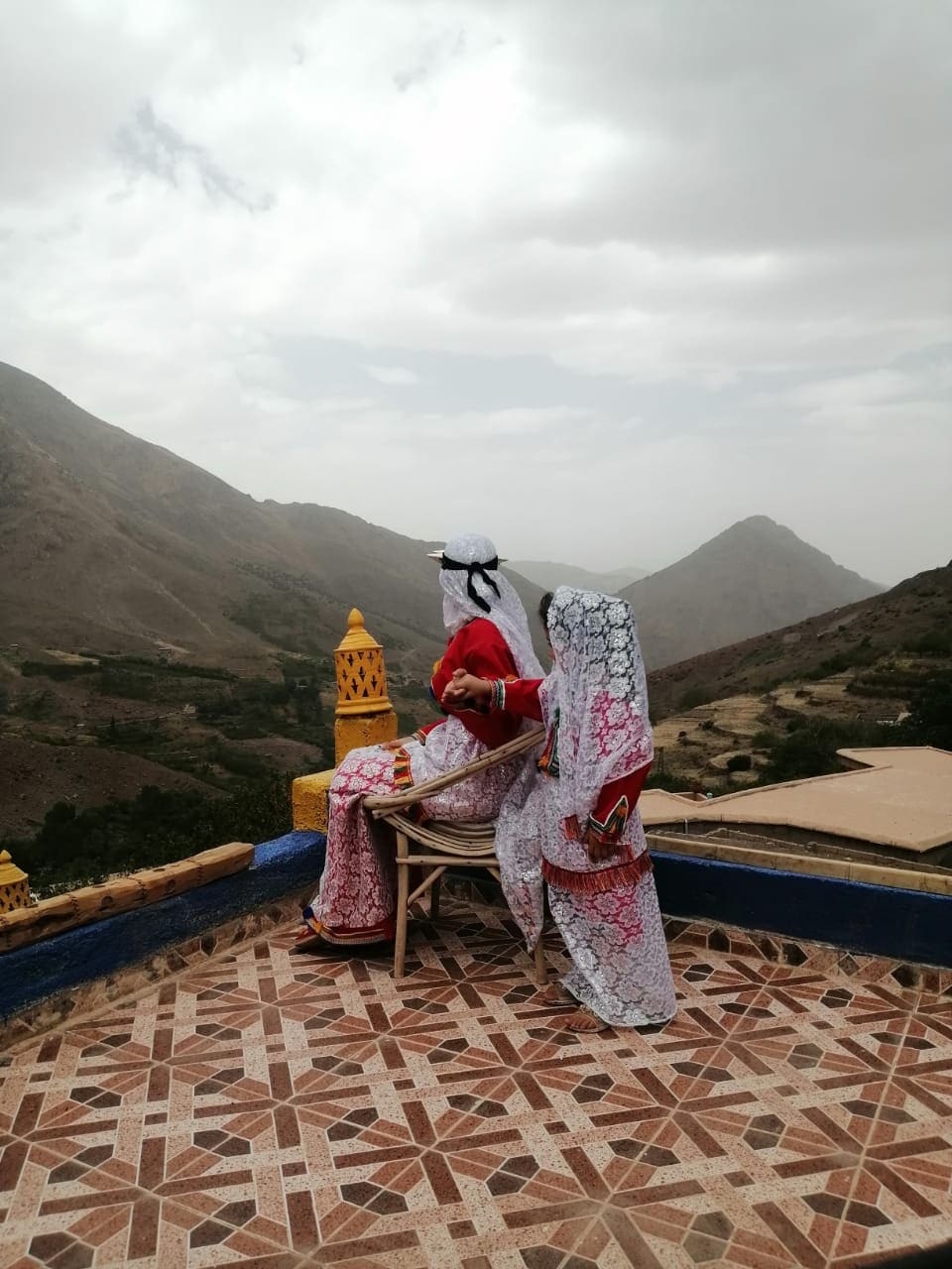 Two women in traditional attire sit on a chair, overlooking a mountainous landscape. The scene includes colorful architectural details, with vibrant tiles and a tower in the background. Soft clouds cover the sky, creating a serene atmosphere.