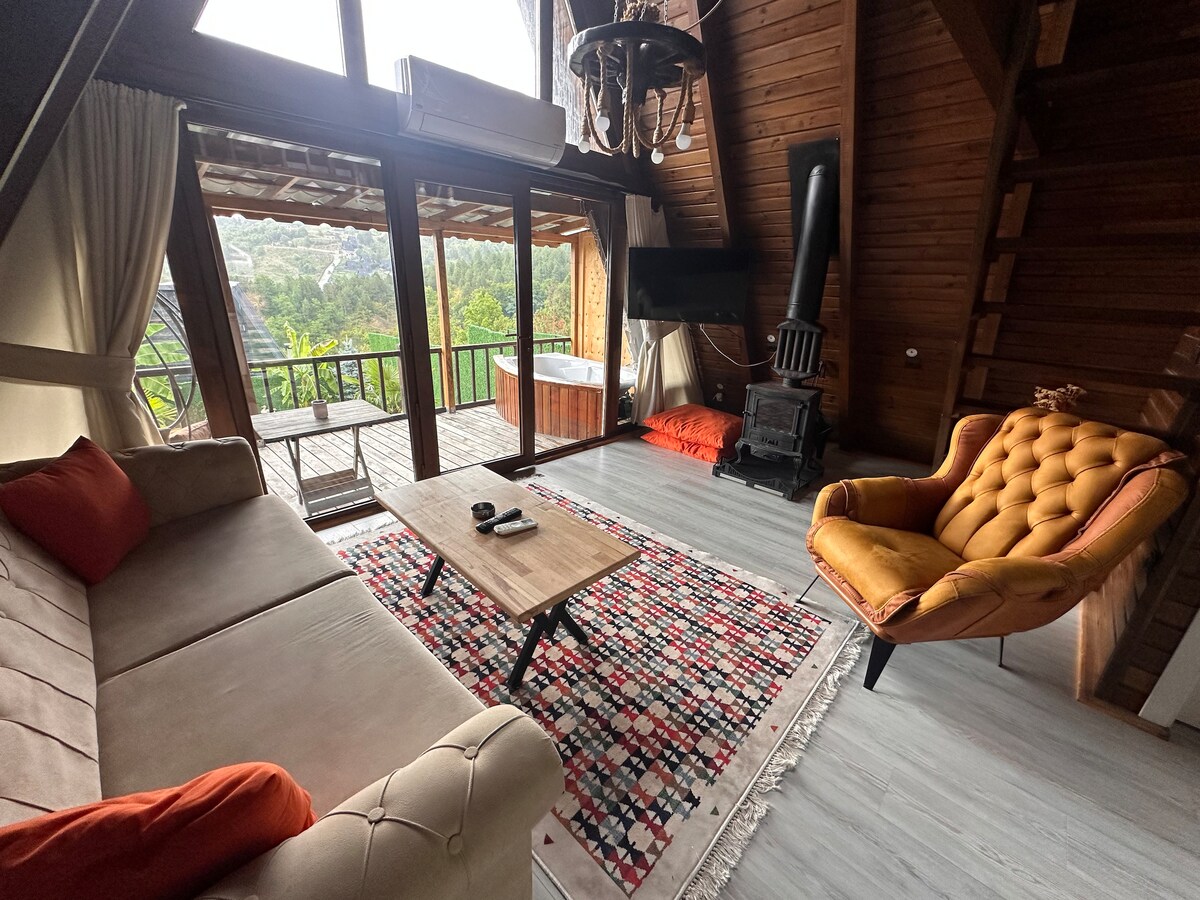 A cozy living area is designed with wooden beams and large windows that provide views of the surrounding nature. A plush sofa and an armchair are positioned around a coffee table, with a colorful patterned rug beneath. A hot tub is visible on the balcony.