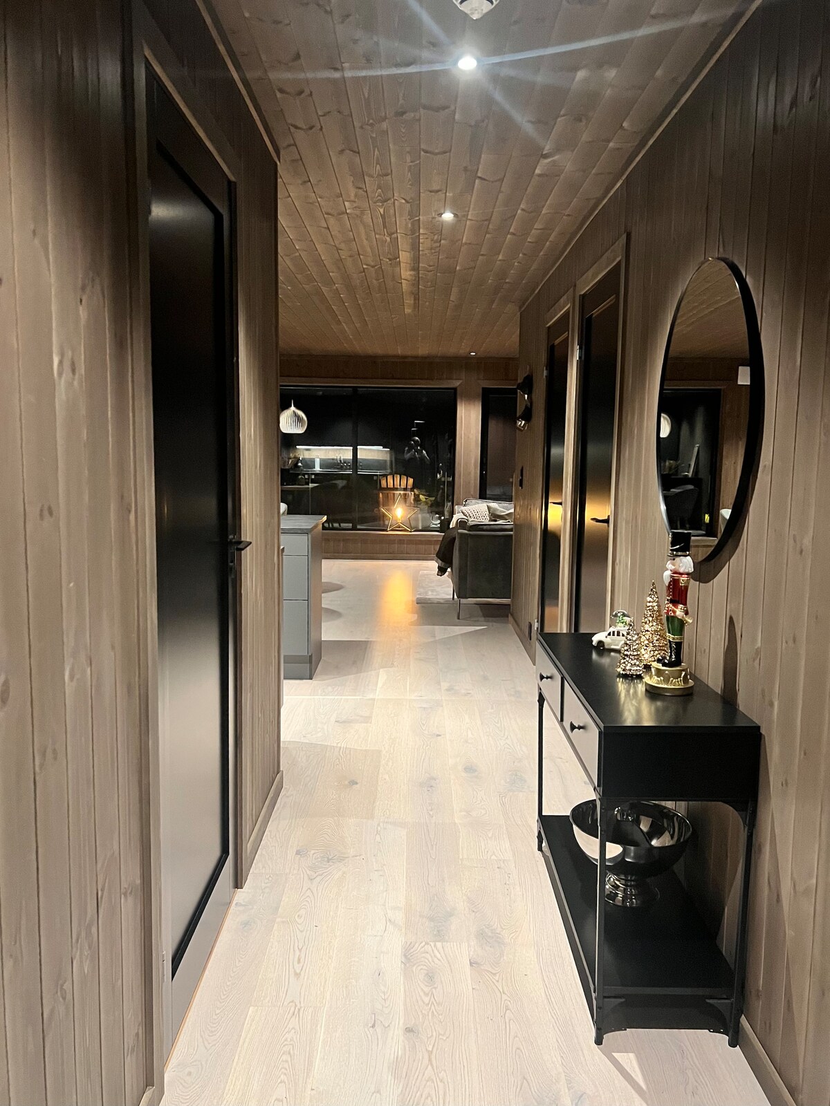 A well-lit hallway is lined with warm wooden paneling, leading towards an open living area. A round mirror reflects the space, while a black console table with decorative items is positioned along one wall. Soft lighting creates a cozy atmosphere.