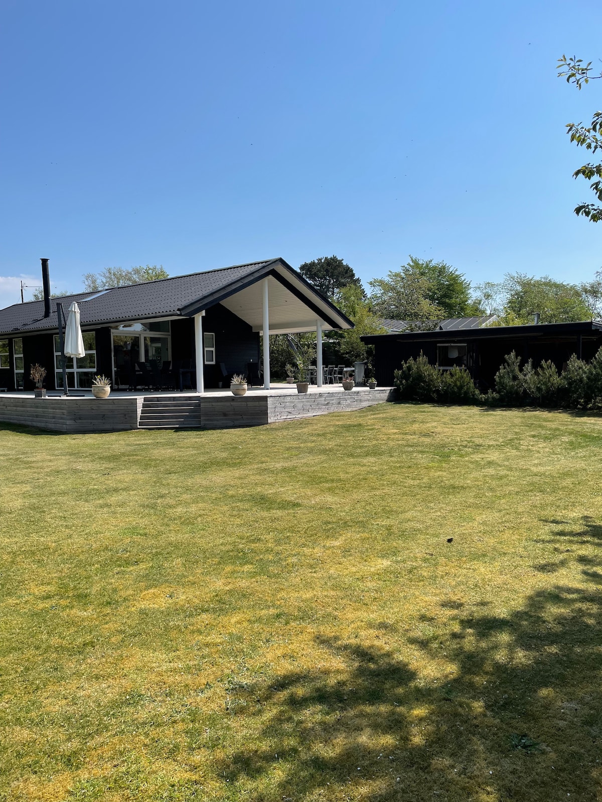 A renovated summer house is set in a spacious yard, surrounded by greenery. The structure features a welcoming porch with steps leading up and a patio umbrella. A second building is visible in the background, enhancing the property’s outdoor space.