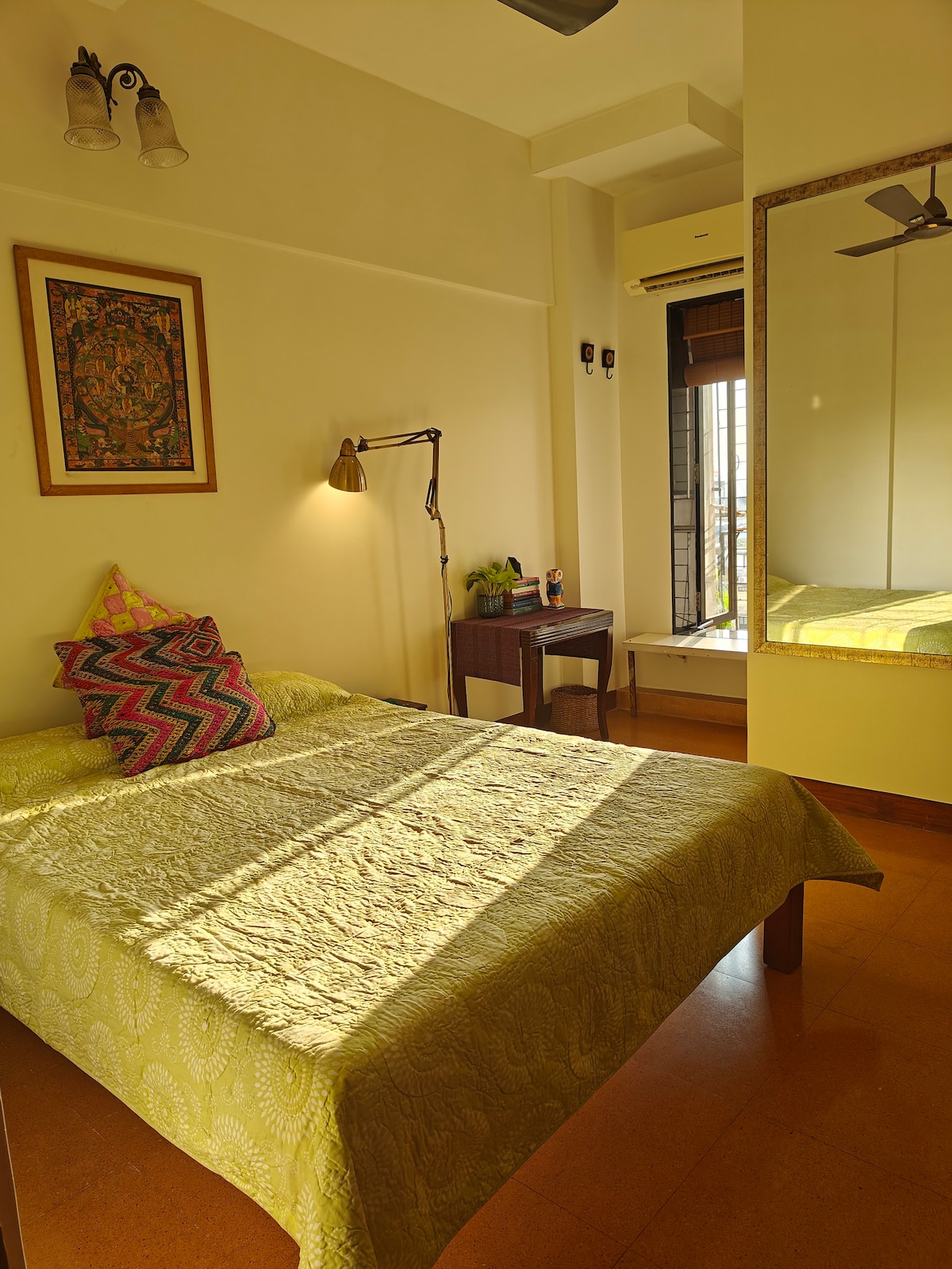 A cozy bedroom featuring a spacious bed topped with a green quilt. A bedside lamp illuminates a corner, enhancing the simple decor. A desk is positioned near a window, with sunlight casting shadows across the room. A mirror reflects part of the space, adding depth.