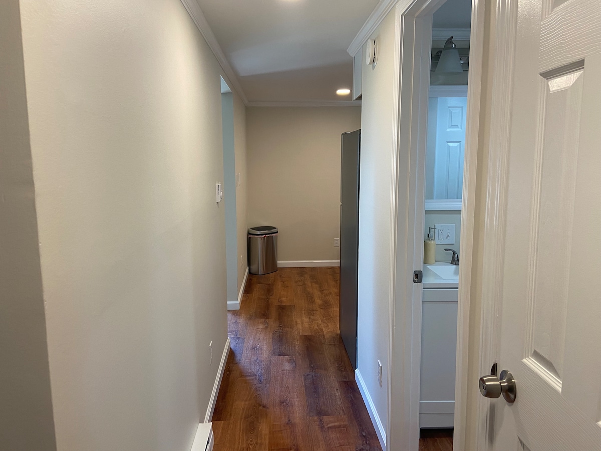 A hallway with a clean, neutral-colored wall and warm wooden flooring is visible. A doorway on the right leads to a bathroom area, while another room is noted at the end of the hallway. A trash bin is situated to the left.