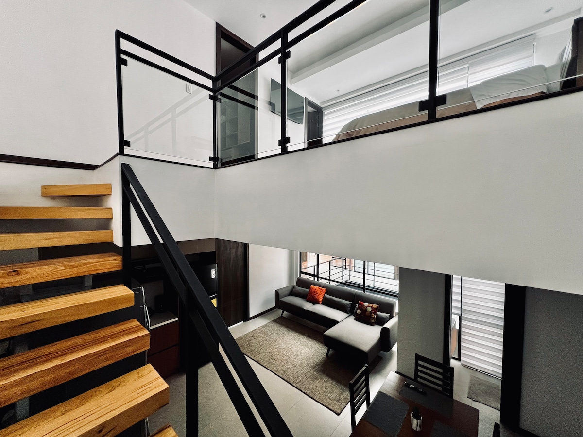 A two-level loft is featured, with a wooden staircase leading to the upper sleeping area. Large windows provide natural light to the open living space, which includes a comfortable sofa and a dining area. A neutral color palette creates a modern ambiance.