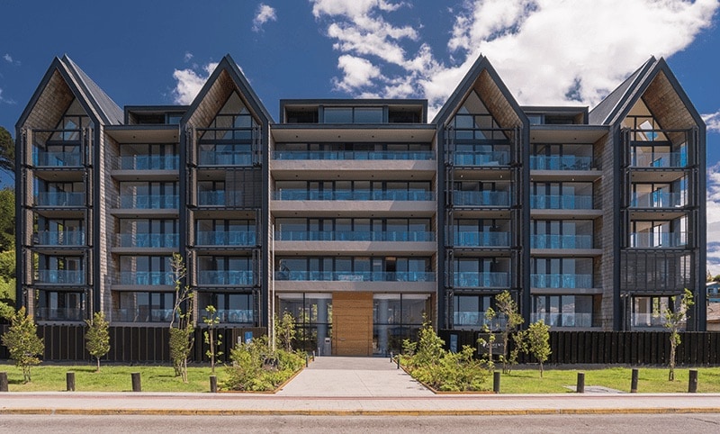 The modern building features a distinct architectural design with multiple balconies and large glass windows, allowing ample natural light. Lush greenery is seen surrounding the entrance, which is accompanied by a neatly paved pathway leading to the welcoming entrance.