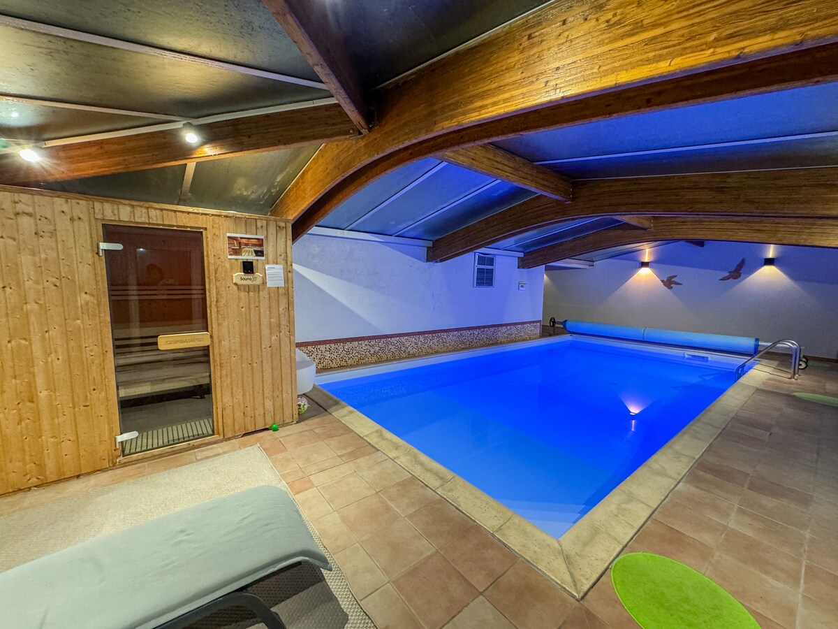An indoor heated pool is framed by wooden beams and includes a sauna area. The pool is illuminated in soft blue light, creating a tranquil atmosphere. A lounged chair sits nearby, and the tiled floor adds a touch of warmth to the space.