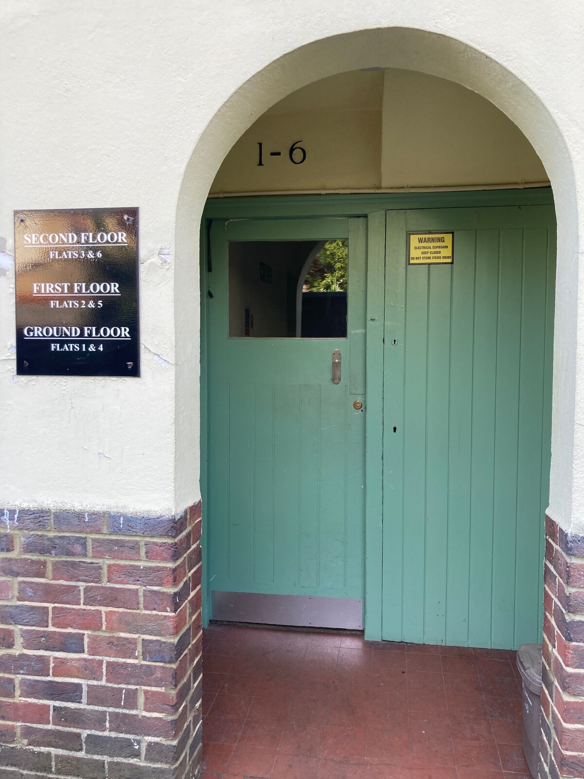 An entrance area is visible, featuring an archway and a green door. A plaque indicating the floor numbers and flat numbers is mounted on the wall to the left. The flooring is tiled in a reddish hue, and light filtered through the doorway creates a welcoming atmosphere.