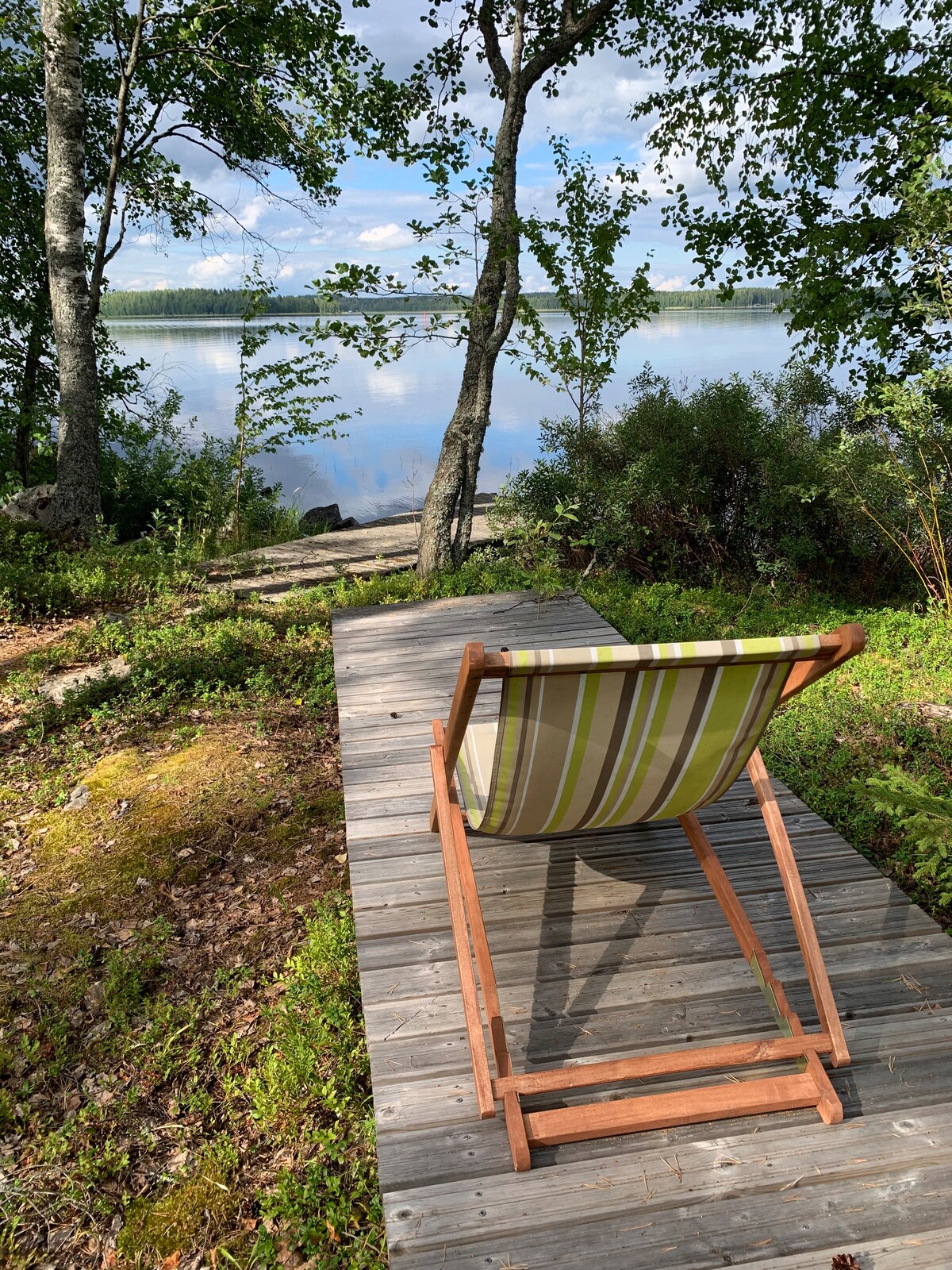 A wooden deck extends over a tranquil lake, bordered by lush greenery. A striped lounge chair is positioned to face the calm water, framed by trees and gentle hills in the background. Soft sunlight casts a serene ambiance on the scene.