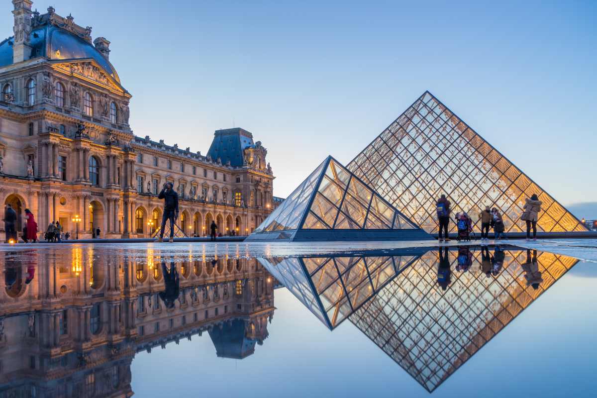 The Louvre Museum showcases its iconic glass pyramid and historic architecture at sunset. The glass structure reflects the warm colors of the sky and the museum's ornate façade, providing a captivating view. Visitors can be seen enjoying the space, enhancing the atmosphere of cultural appreciation.