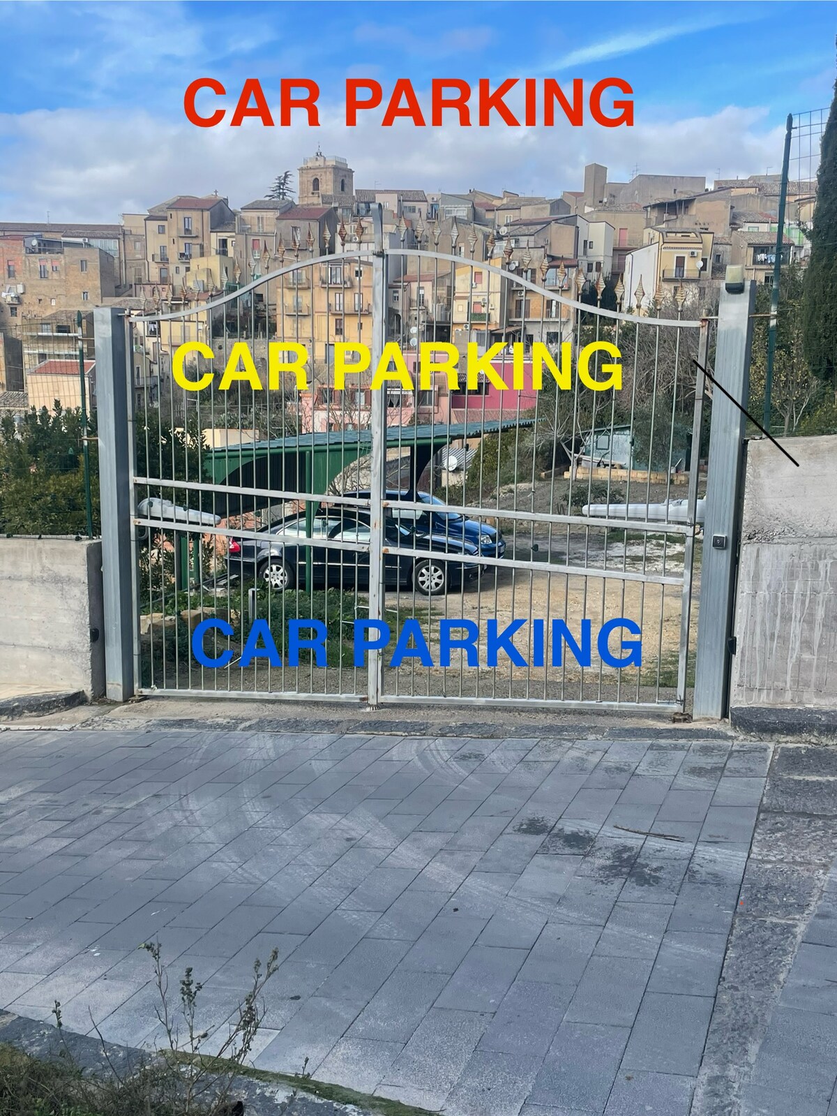 A secure parking area is depicted, featuring a gated entrance that opens to a designated parking space for vehicles. The surrounding scenery includes a view of the hillside town, with buildings visible in the background.