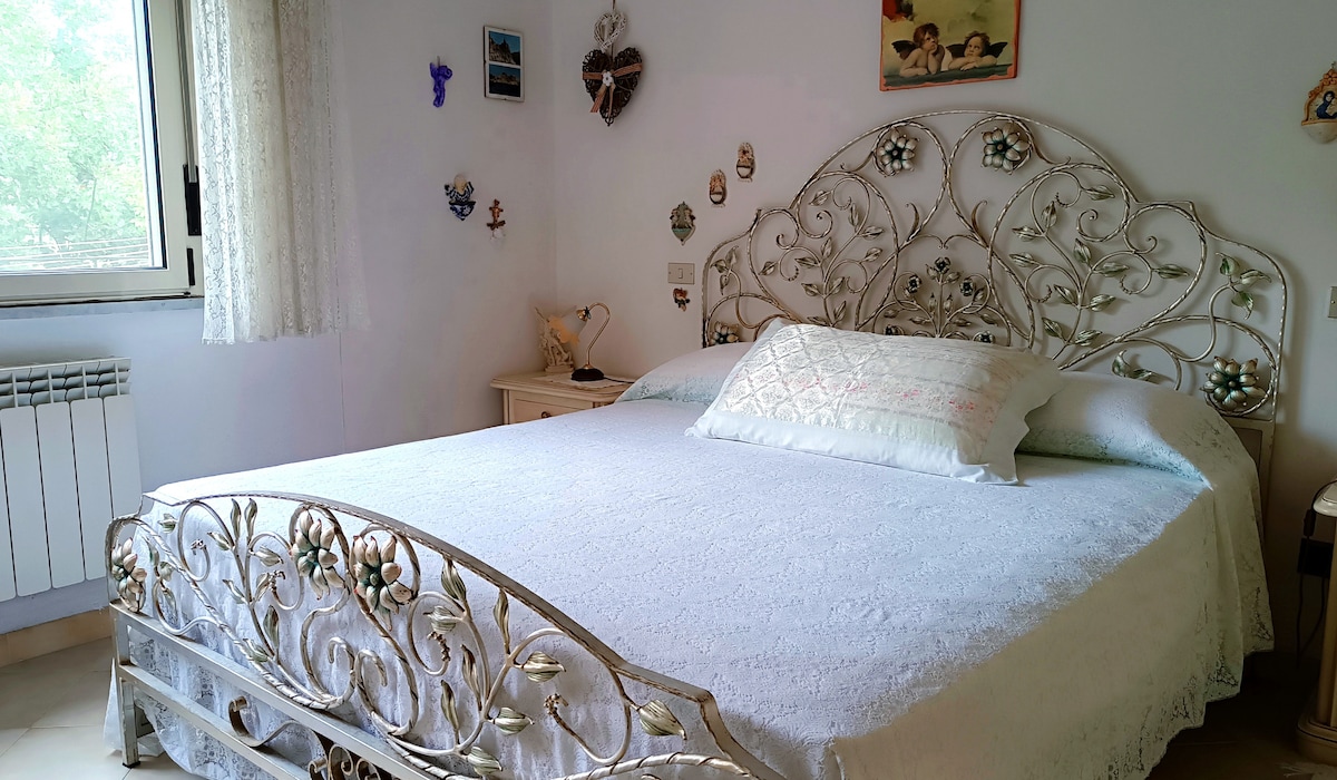 A spacious bedroom features an ornate metallic bed with a white embroidered bedspread. The walls are decorated with various wall art and small decorative objects. Natural light filters through a window, enhancing the calming environment.