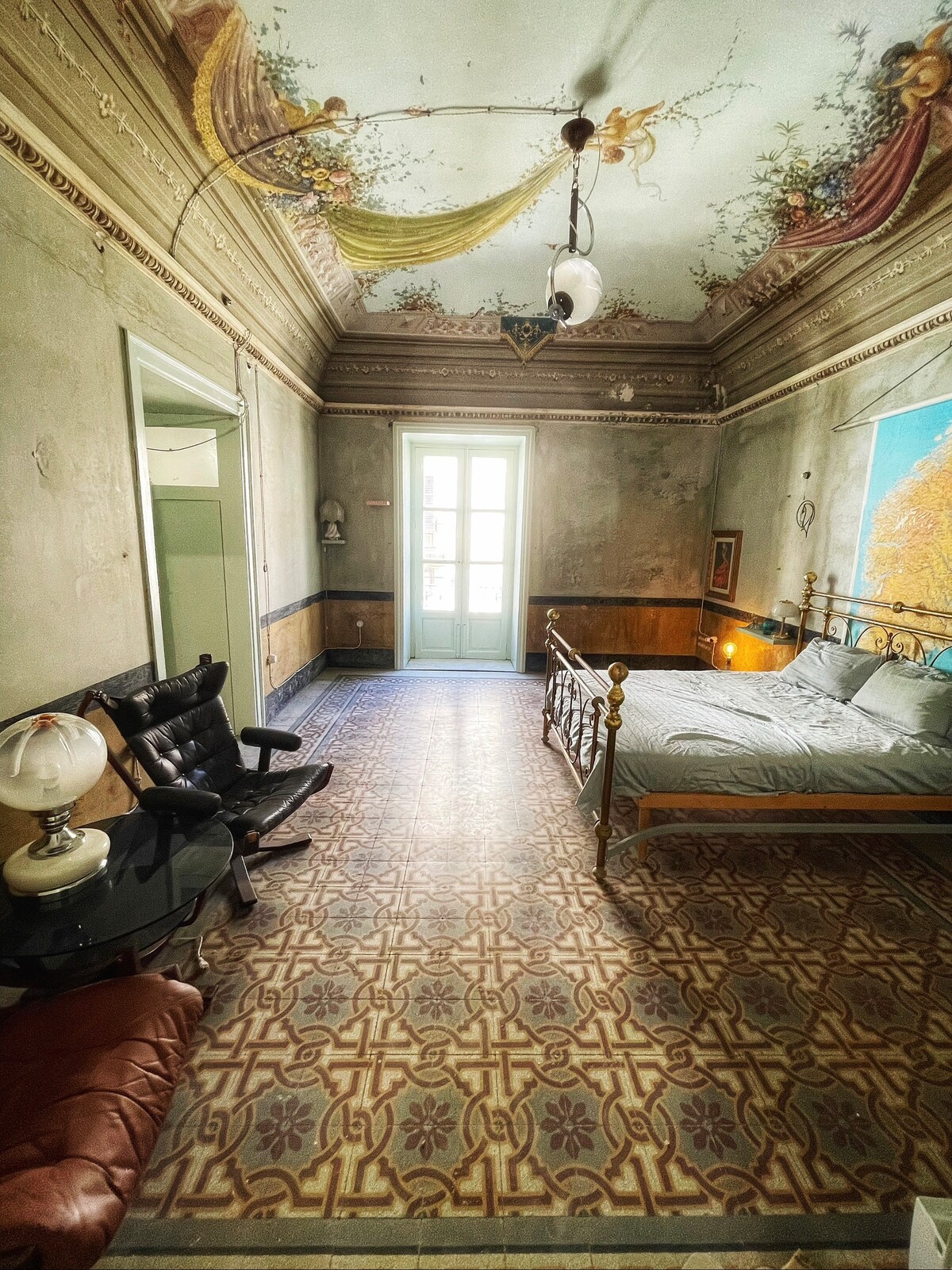 A bedroom is showcased with a vintage atmosphere, featuring a spacious bed with light bedding and elegant ornamental details on the ceiling. Sunlight enters through double doors, illuminating beautifully patterned tiles on the floor. Antique furniture, including a lamp and armchair, enhances the room's charm.