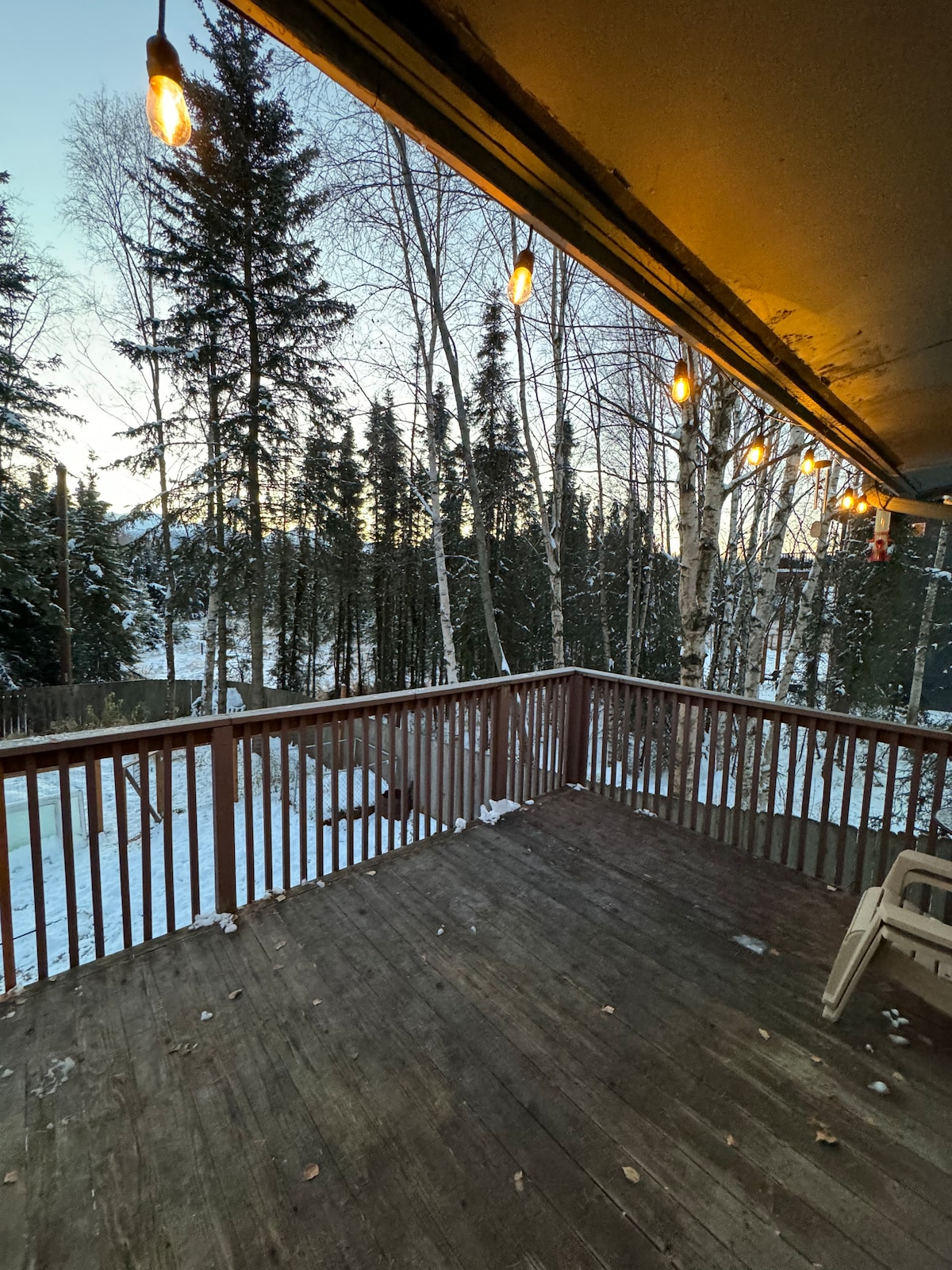 A spacious wooden deck is visible, surrounded by tall trees and gently falling snow. String lights hang above, providing soft illumination. The ground is covered in snow, creating a serene winter setting that invites outdoor relaxation.