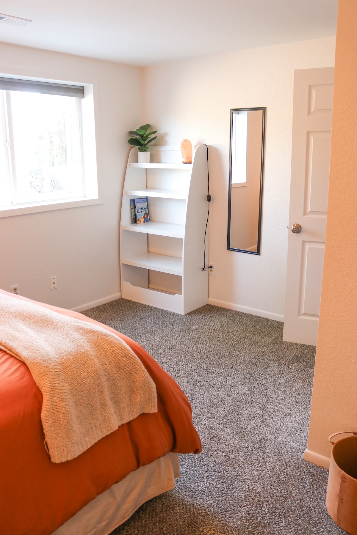 A cozy bedroom features a spacious bed with an orange duvet and a soft cream throw. Natural light enters through a window beside a simple bookshelf and a floor mirror. The carpeted floor enhances the comfort of the room.