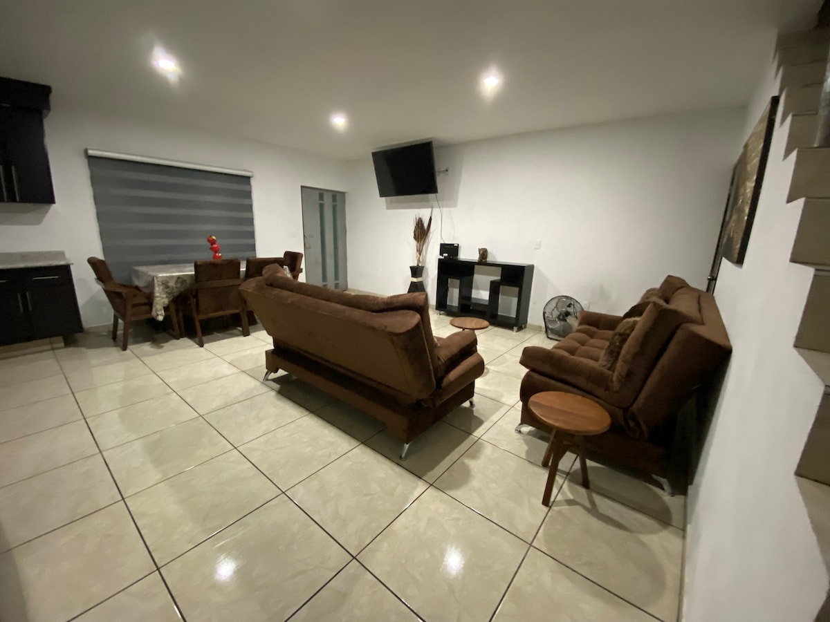 A spacious living area is presented, featuring two brown sofas facing each other. A small wooden side table is placed beside one sofa. A dining table is located in the corner, with four chairs arranged around it. Light tiles enhance the room's brightness.