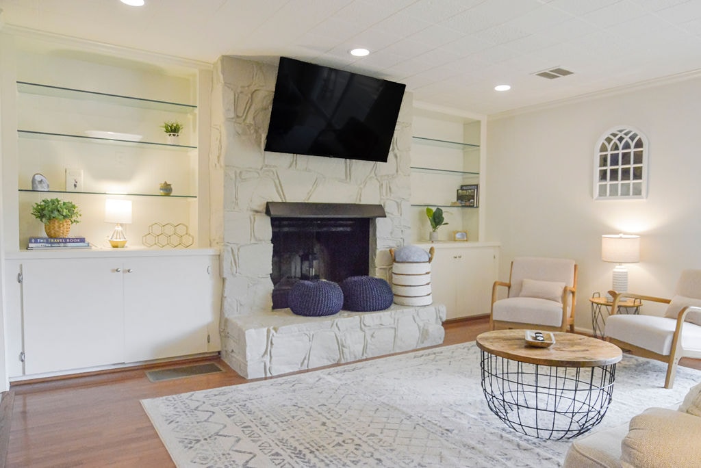 A cozy living area features a stone fireplace with a mounted flat-screen TV above. Two comfortable chairs are positioned beside a wooden coffee table, which rests on a patterned area rug. Shelving units display decorative plants and items, enhancing the inviting atmosphere.