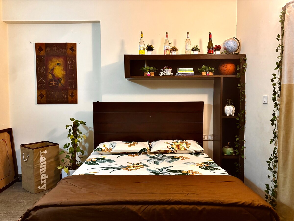 A cozy sleeping area is presented with a spacious bed adorned with floral patterned bedding. A wooden shelf above showcases decorative items and plants, while a painting hangs on the wall. A laundry basket is partially visible to the side, adding functionality to the space.