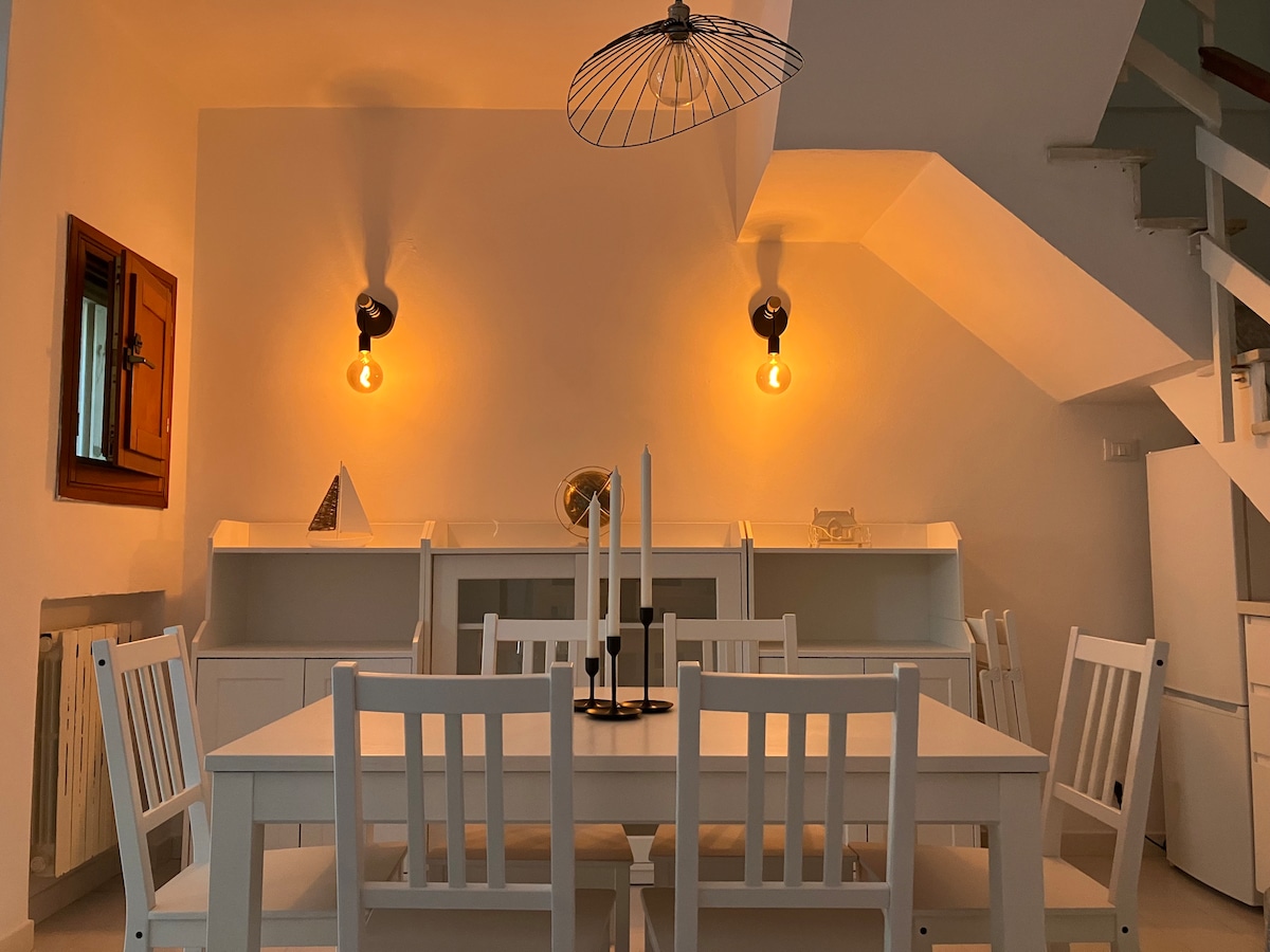 The dining area features a white table surrounded by six matching chairs. Warm light is provided by wall fixtures and a pendant lamp, creating a cozy ambiance. A staircase is visible in the background, along with a built-in shelf and decorative elements.