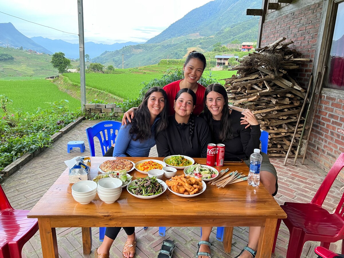 A wooden table is set outdoors, surrounded by individuals enjoying a variety of dishes. Colorful bowls of food, including salads and fried items, are arranged alongside beverages. Lush green hills and distant structures can be seen in the background, adding to the tranquil setting.