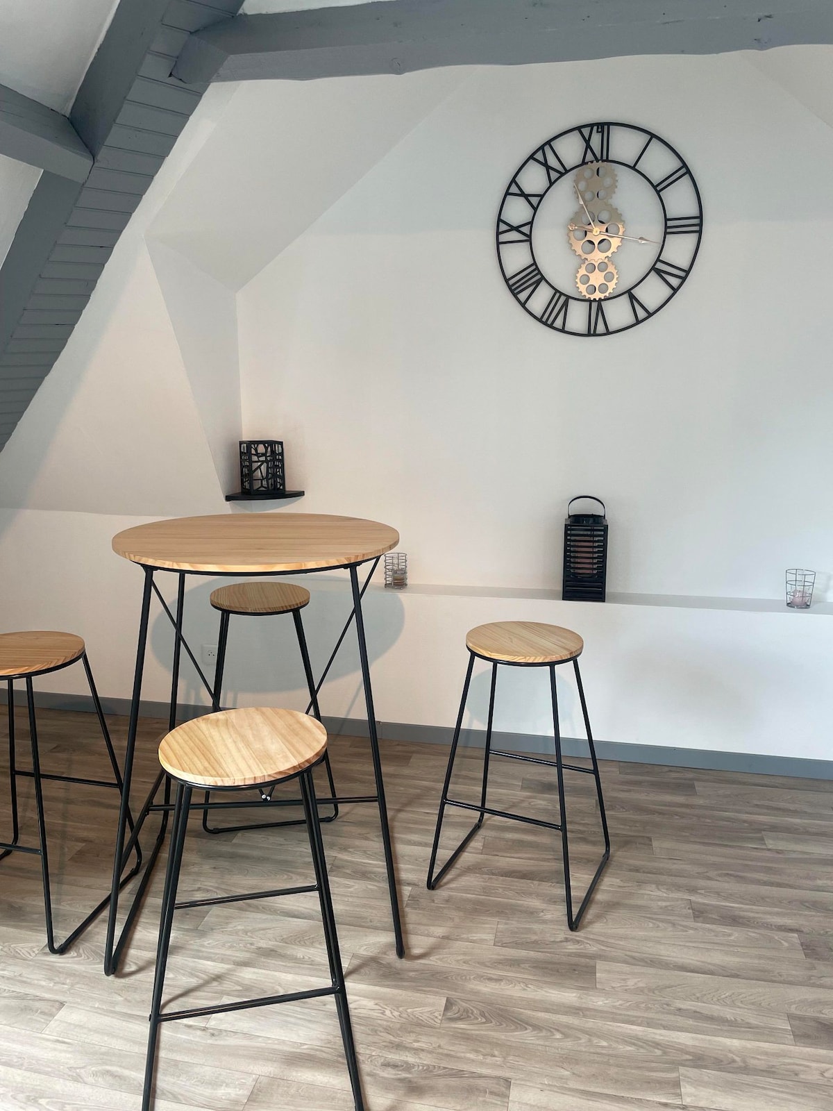 A minimalist dining area features a circular wooden table accompanied by four metal bar stools. The light wood surfaces contrast with the dark metal, while a stylish wall clock and lanterns add a decorative touch against the white wall.