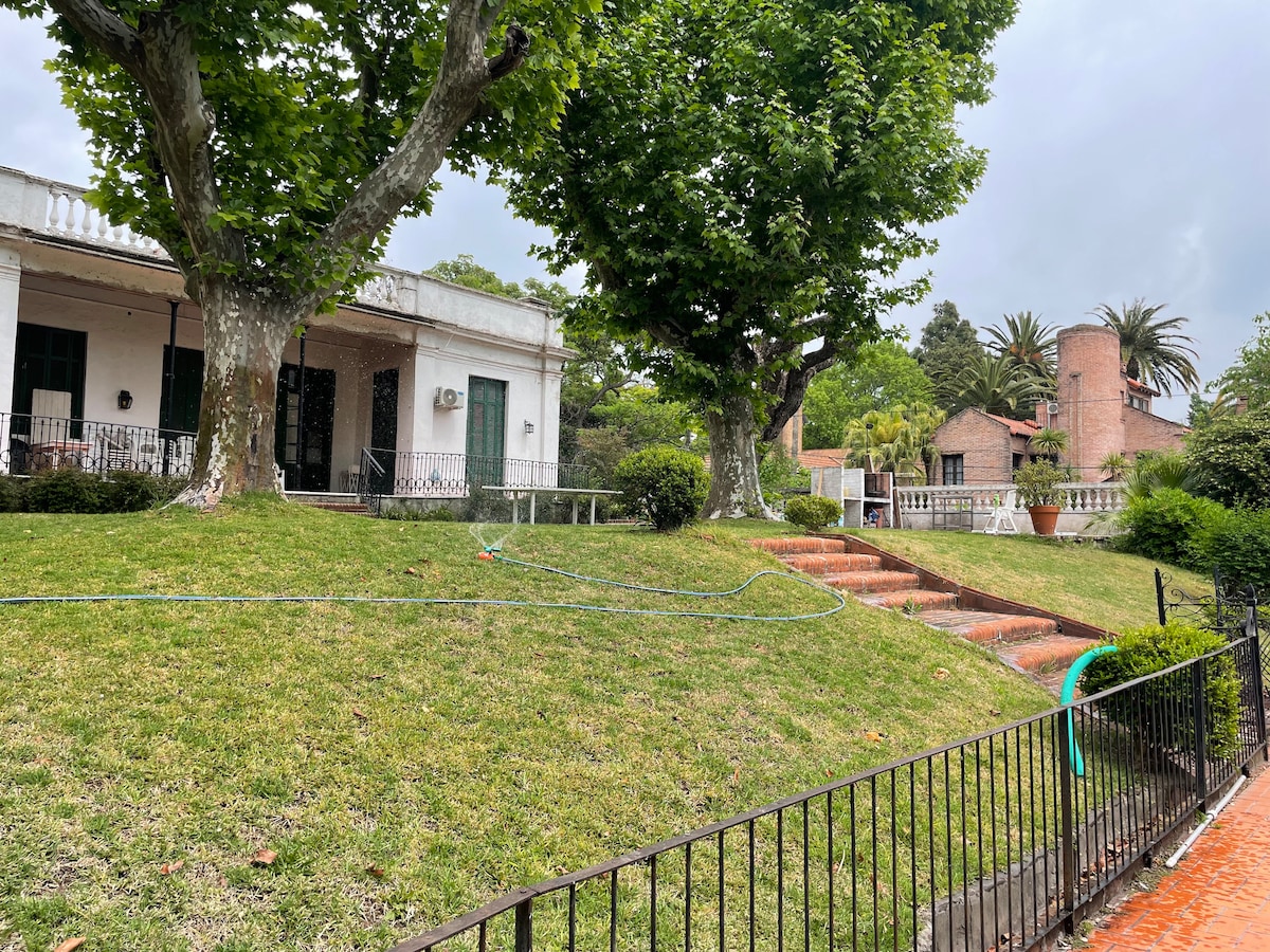 A well-maintained garden is framed by large trees, offering a green and inviting space. A set of stone steps leads from the garden to the entrance of the colonial house. The exterior features a white façade with green doors and windows.