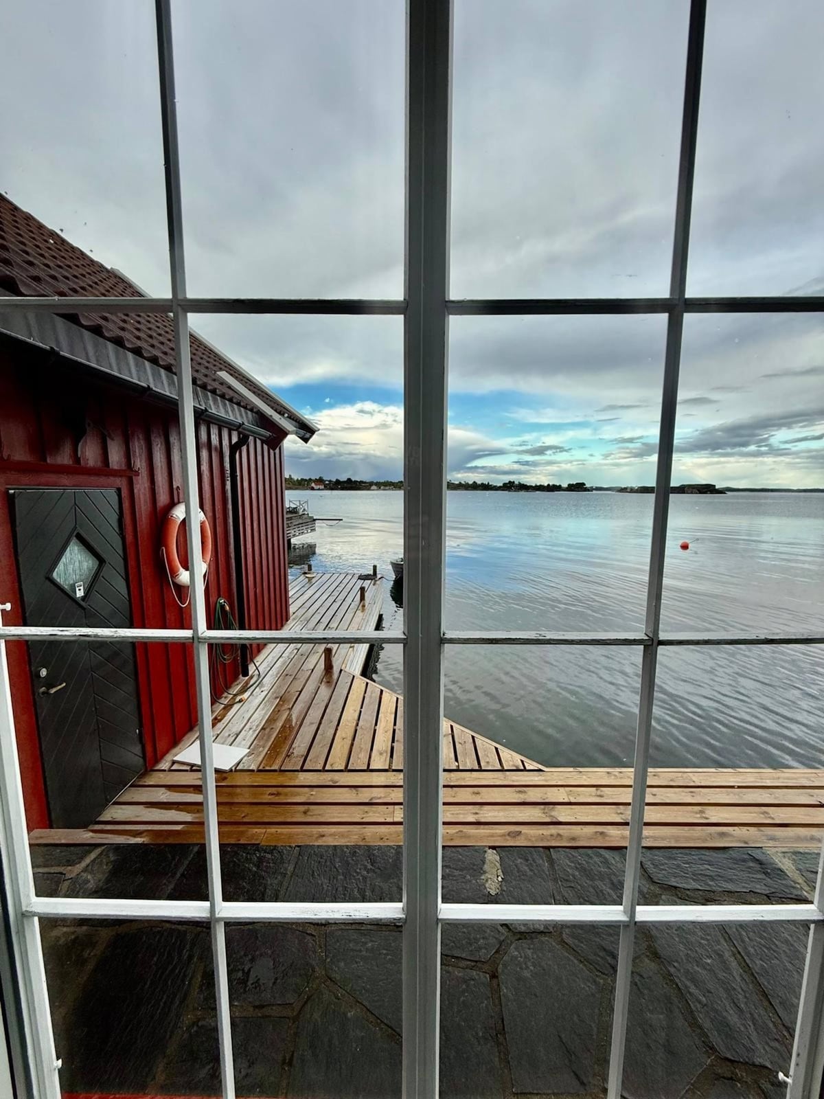 A view from a window reveals a peaceful waterside scene. A wooden jetty extends into calm water, with a backdrop of gentle clouds and distant land. A red cabin is visible nearby, adding a charming element to the setting.