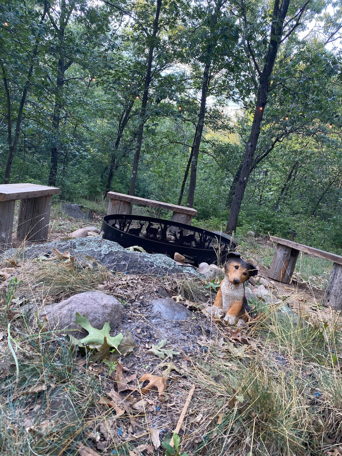 An outdoor firepit is surrounded by natural stone and rustic wood seating, with the forest backdrop providing a serene ambiance. Fallen leaves and grasses cover the ground, while a small, decorative dog figure adds a playful touch to the campsite area.