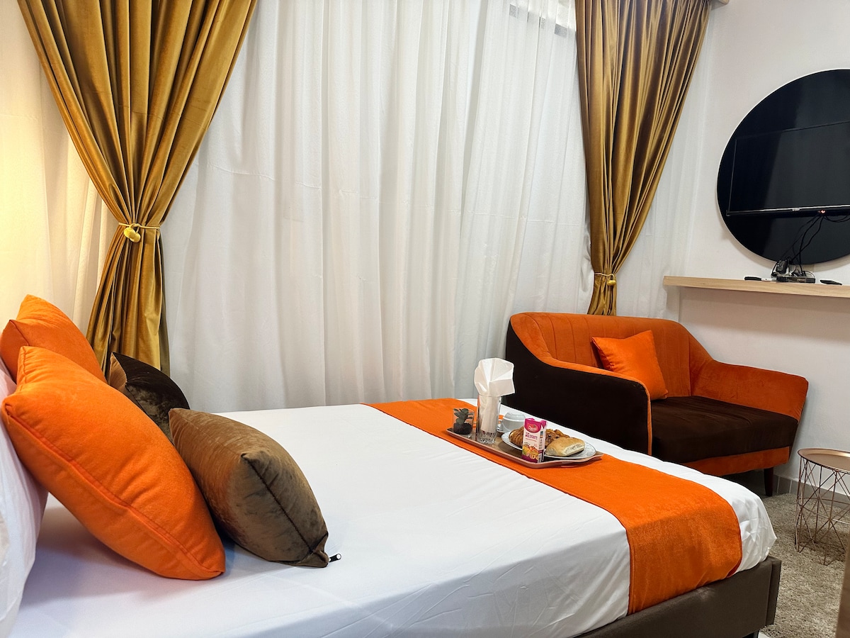 A comfortable sleeping area is presented with a spacious bed featuring an orange and white runner. Soft, textured pillows are arranged atop the bed. A modern television is mounted on the wall above a sleek shelf. An inviting seating area includes an orange armchair and a small table.