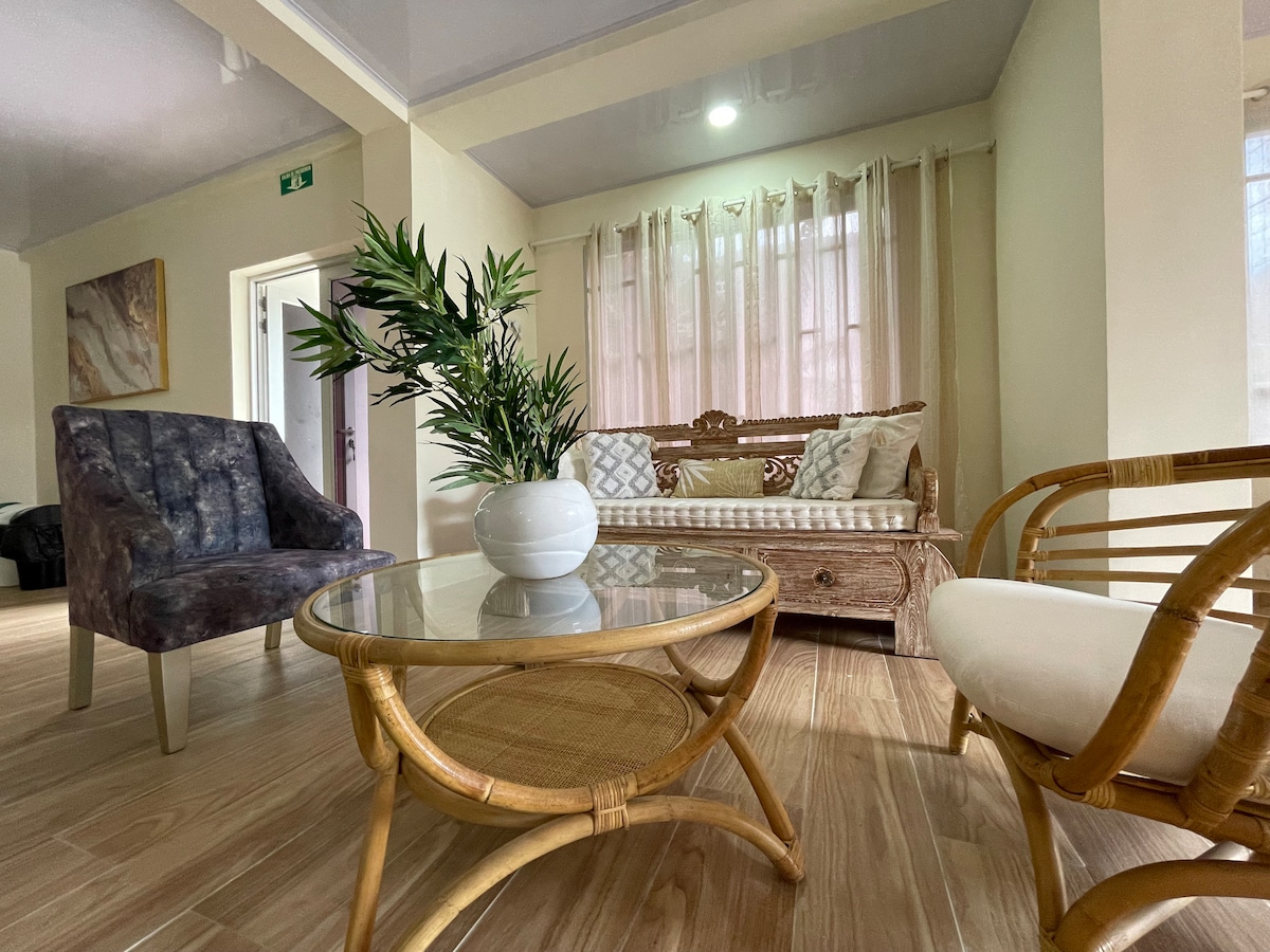 A softly lit living area features a comfortable sofa along with a plush armchair. A round glass table rests in the center, accompanied by a decorative indoor plant. Sheer curtains allow natural light to filter through, enhancing the open space.