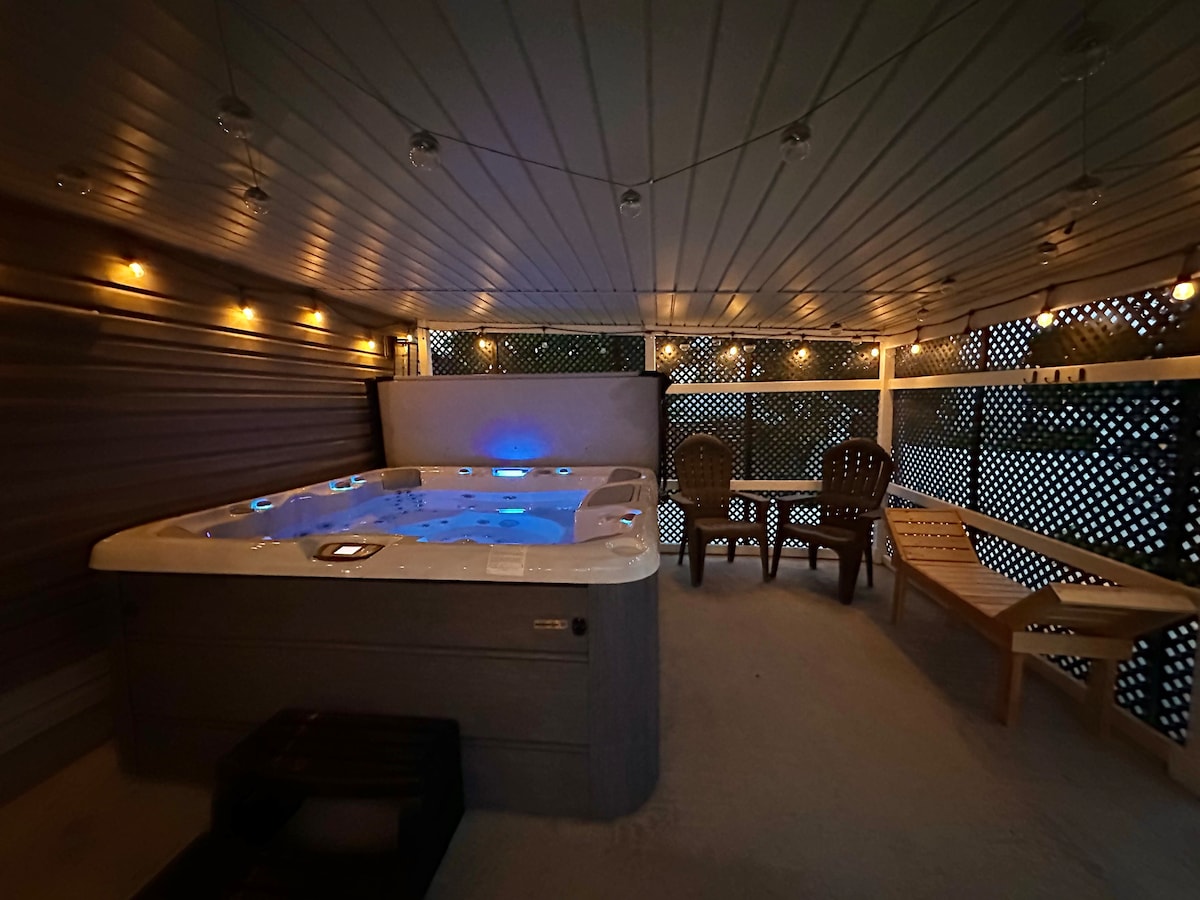 A well-lit hot tub area features a spacious hot tub with blue lighting, surrounded by decorative string lights. Comfortable seating is provided with two chairs and a lounge chair, all set against a backdrop of lattice walls for a sense of privacy.