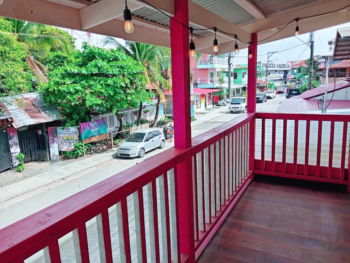 A vibrant balcony is framed with red railings, offering views of the bustling street below. Decorative string lights are hung above, adding a cozy touch. Lush greenery and local businesses are visible in the background, enhancing the lively atmosphere of the town center.