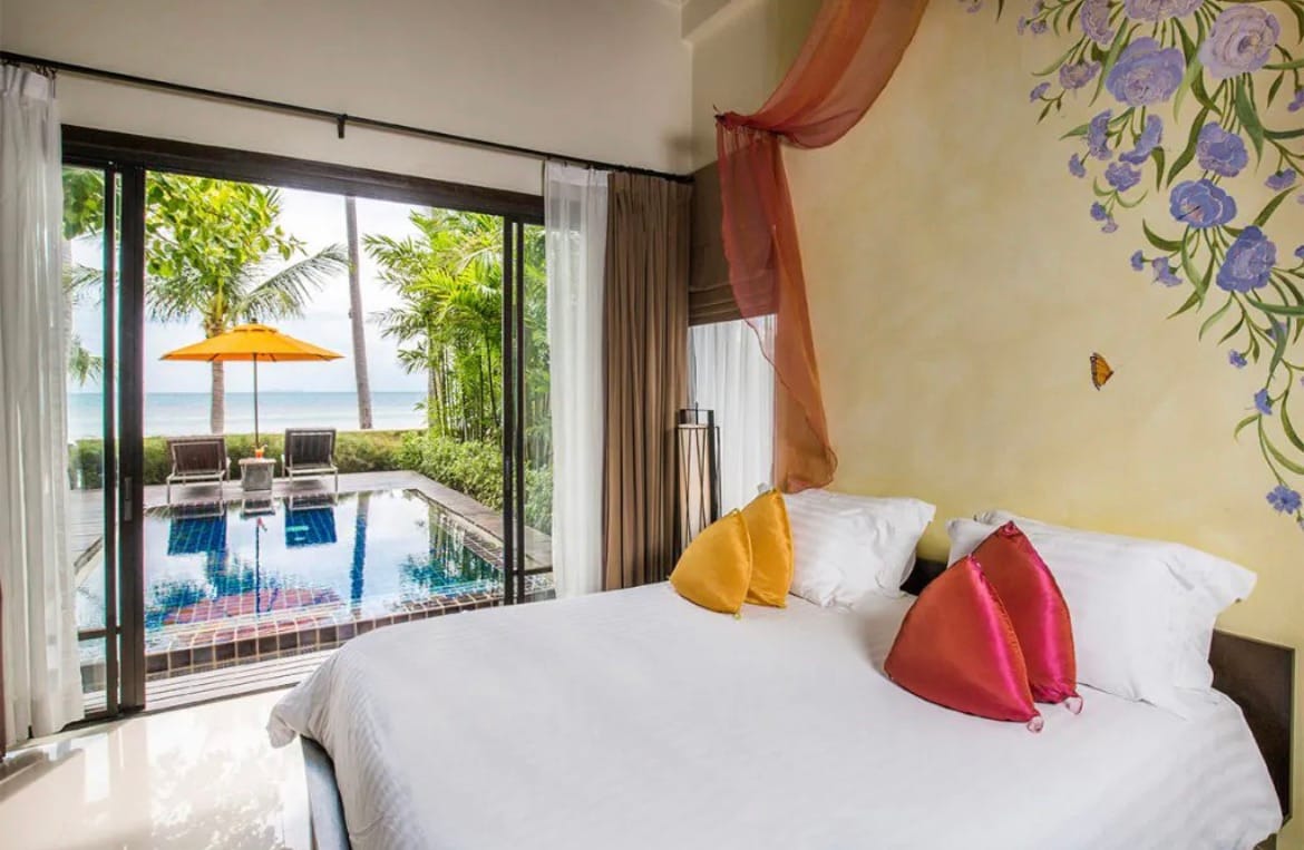 A spacious bedroom features a large bed adorned with colorful pillows. A charming mural with floral accents decorates one wall. Floor-to-ceiling glass doors provide a view of the private pool and beach beyond, framed by palm trees and an orange umbrella.