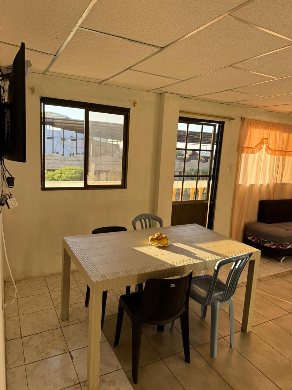 A dining area is presented with a rectangular table surrounded by several chairs in various colors. Natural light is allowed in through two windows, illuminating the space. A television is mounted on the wall, and a comfortable seating area is visible in the background.